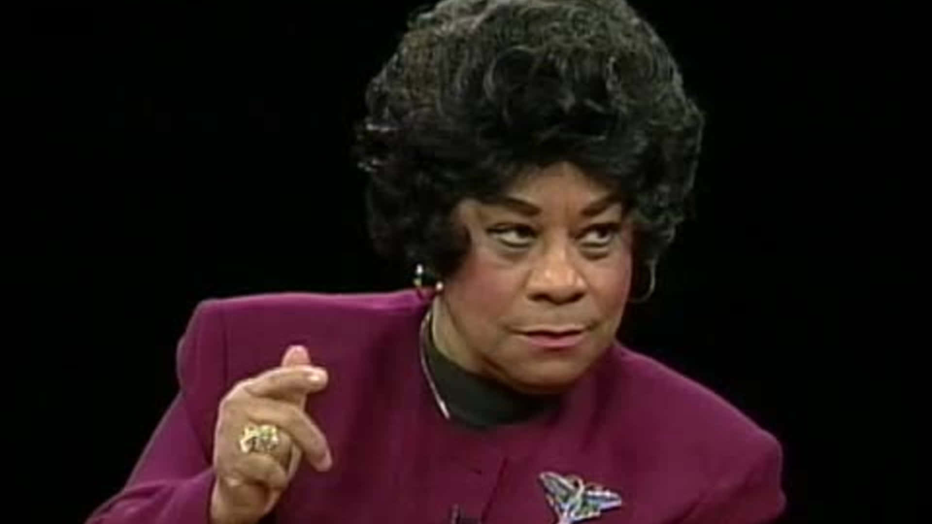 歌手 Ruth Brown 壁紙 — 一位留著捲髮、身穿紫色西裝外套的女性在黑色背景前說話時做手勢。