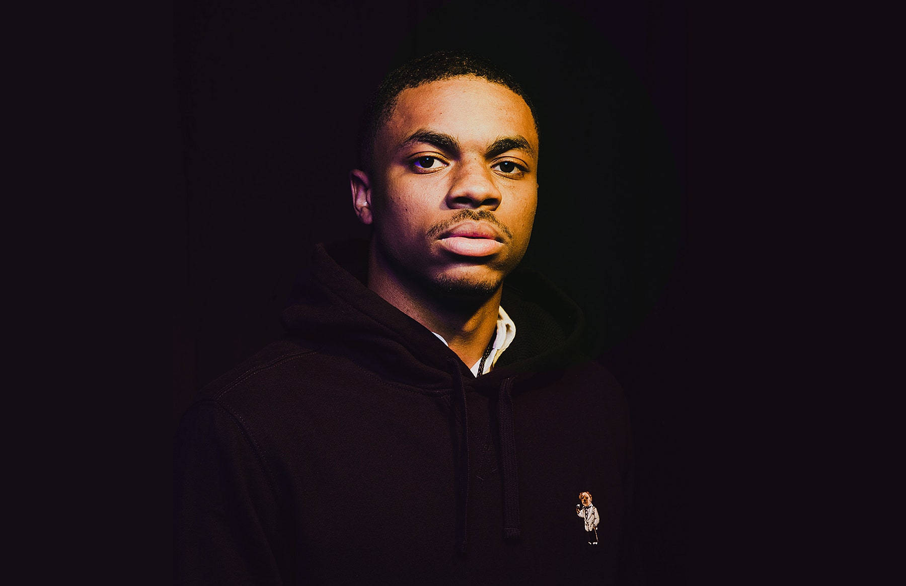 歌手兼饒舌歌手 Vince Staples 壁紙 — 一位穿著深色連帽衫的年輕男子在黑色背景前，於戲劇性的低光照明下直視鏡頭。