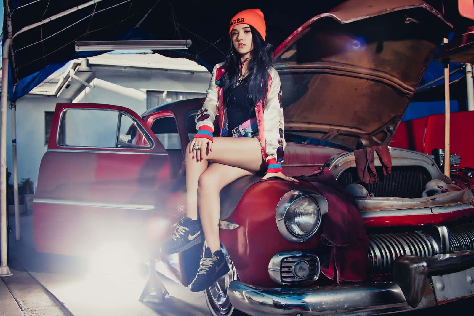 戴橙色冷帽的女性坐在車庫中一輛復古紅色汽車的引擎蓋上，周圍有煙霧。