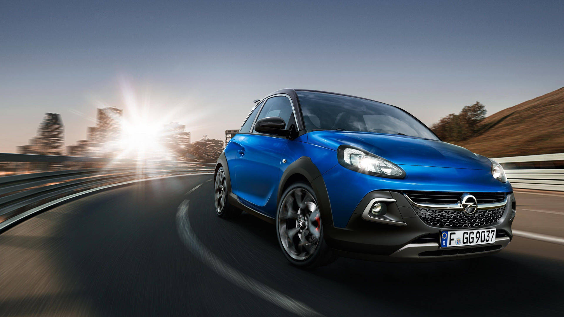 藍色Opel Adam Rocks在日落時分在蜿蜒的道路上行駛，背景是城市天際線。