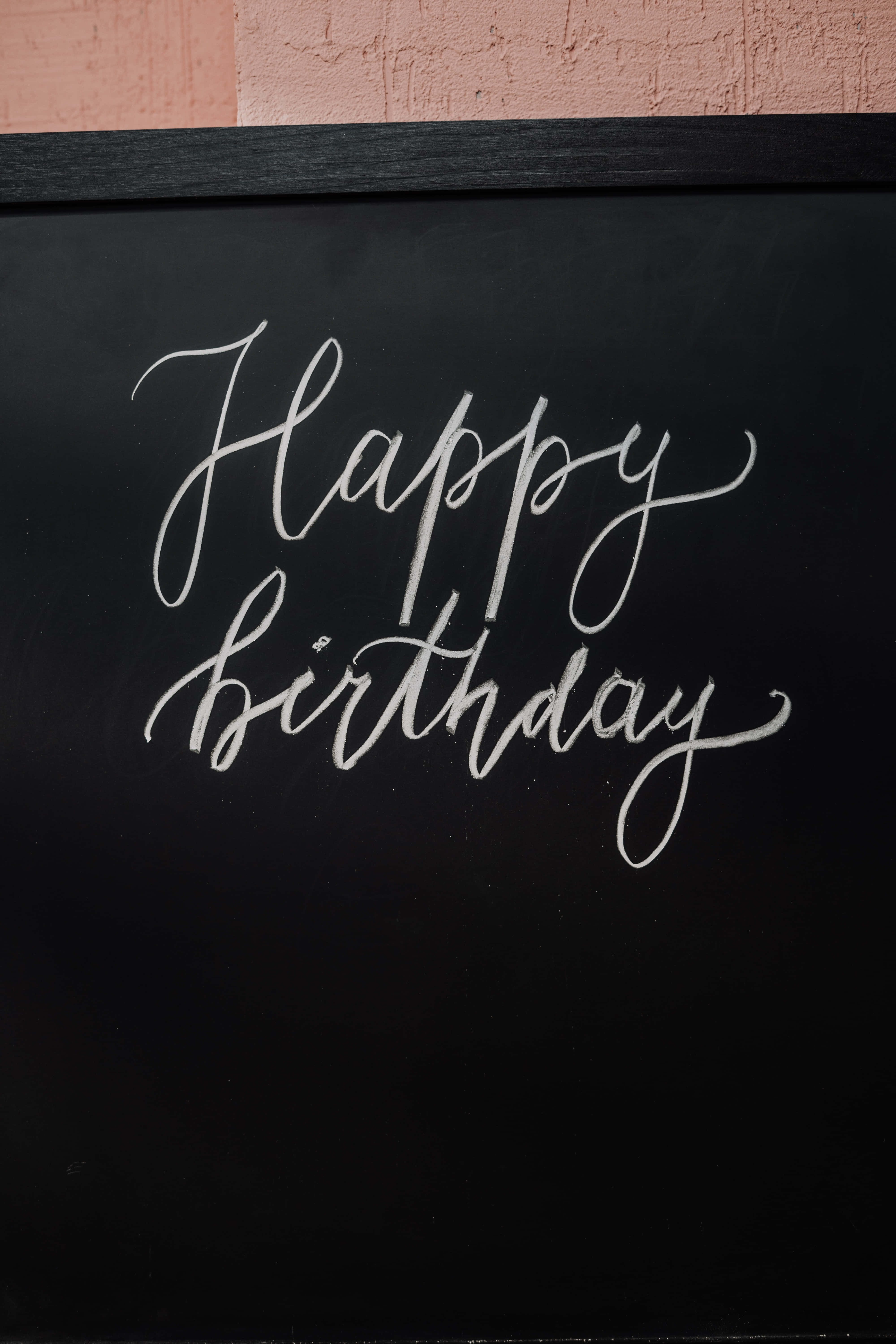 歡慶生日，喜悅無限 壁紙 — 黑色黑板上優雅的白色草書 'Happy Birthday'，上方有紋理的粉紅色牆壁。