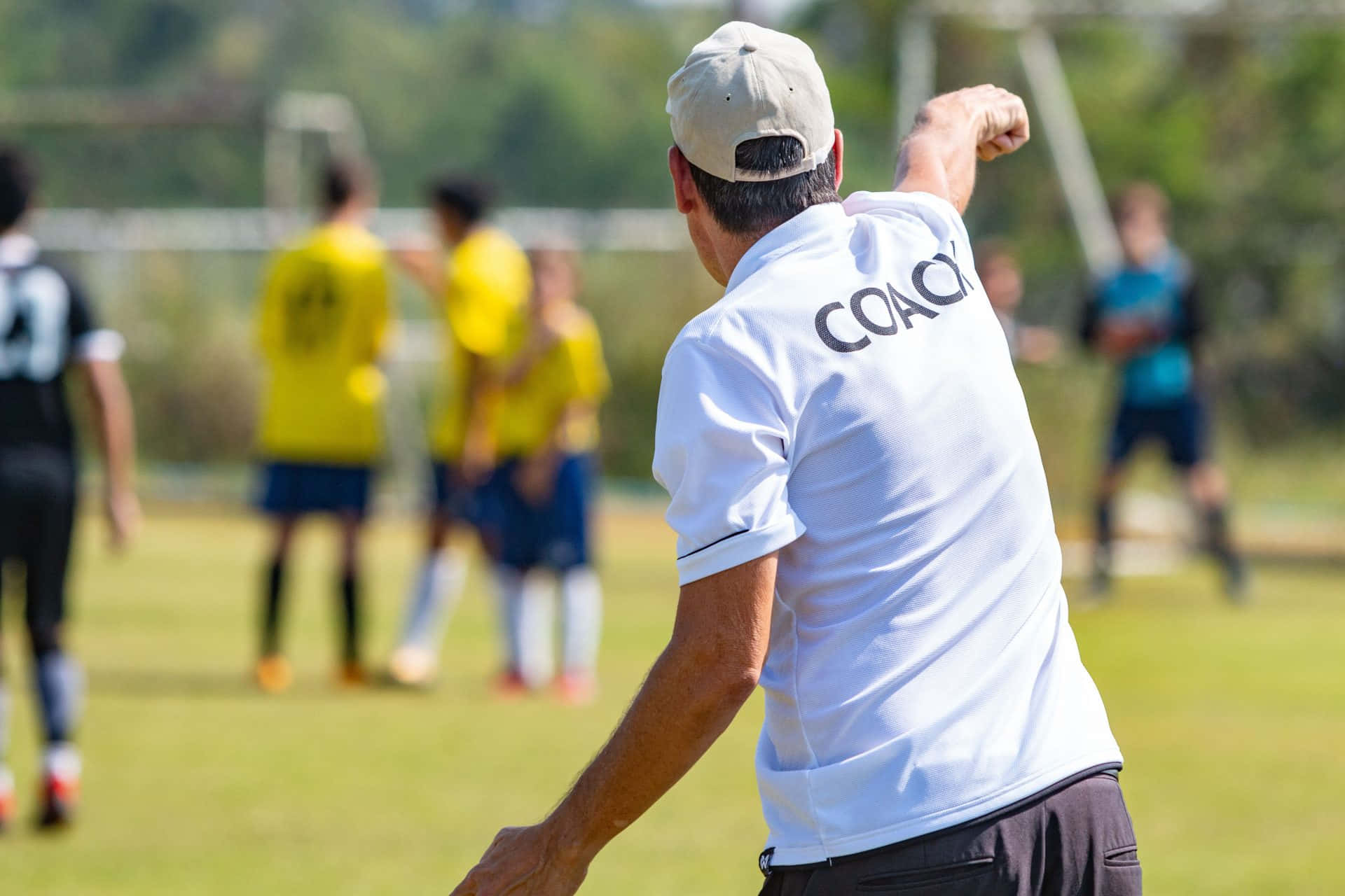 歡樂 Coaching 圖片 壁紙 — 一位身穿白色 'COACH' 上衣的足球教練在陽光明媚的草地上用力做手勢，身後有身穿黃色球衣的球員在訓練。