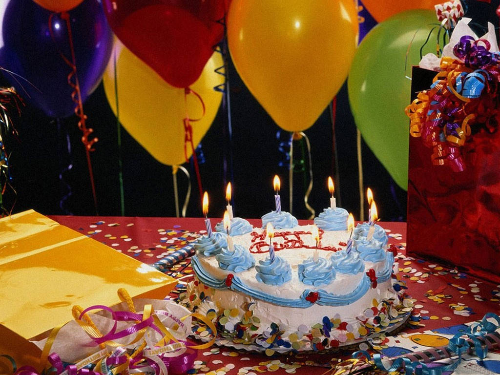 歡樂派對生日蛋糕與蠟燭 壁紙 — 一個插著點燃蠟燭的生日蛋糕放在鋪滿五彩紙屑的桌子上，周圍環繞著彩色氣球和禮物袋。