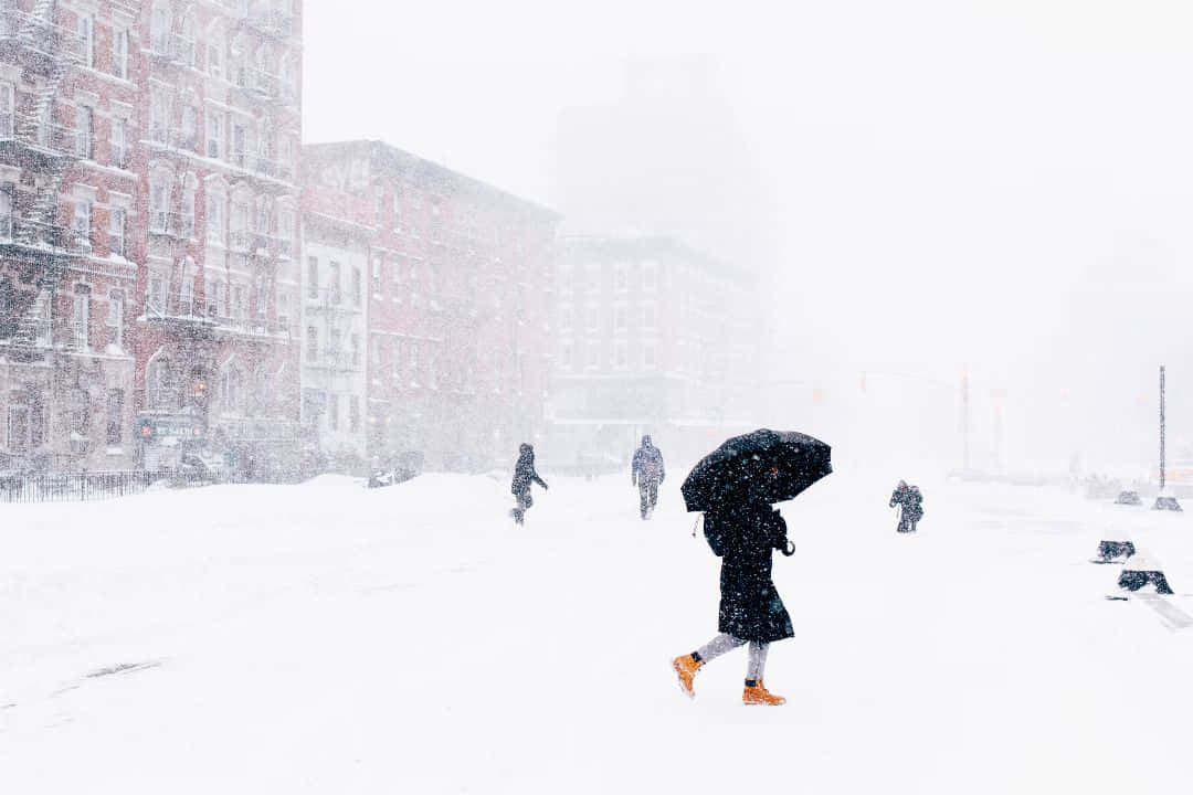 歡樂的暴雪 壁紙 — 一個孤獨的身影穿著黑色外套和橙色靴子，在城市的街道上穿過一場強烈的暴風雪，撐著雨傘，雪花在紅磚建築周圍旋轉。