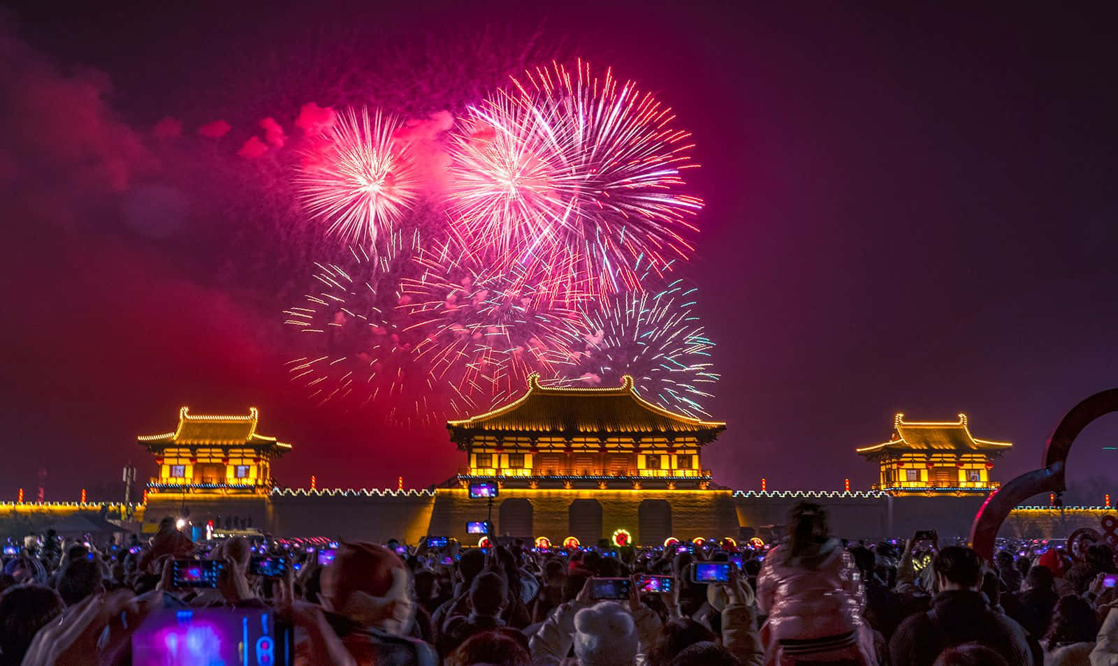 歡樂迎新年 壁紙 — 色彩繽紛的煙花在一個巨大的、燈火通明的古老中國城門上空綻放，人群用手機捕捉這一壯觀景象。