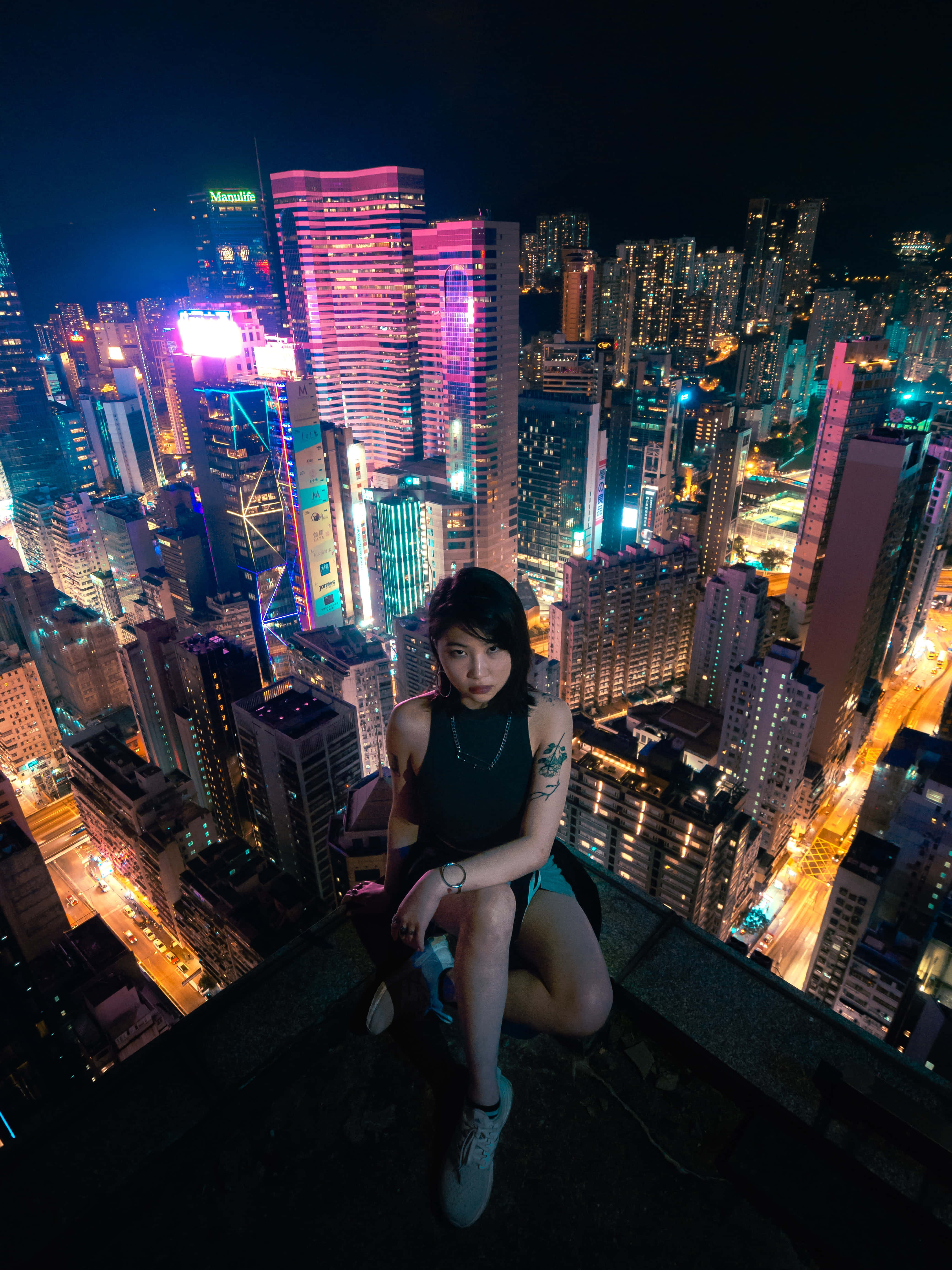 '歡迎來到世界之巔！' 壁紙 — 一位年輕女性在夜晚坐在摩天大樓頂端，俯瞰著耀眼的霓虹燈城市景觀，高聳的建築物和發光的街道。