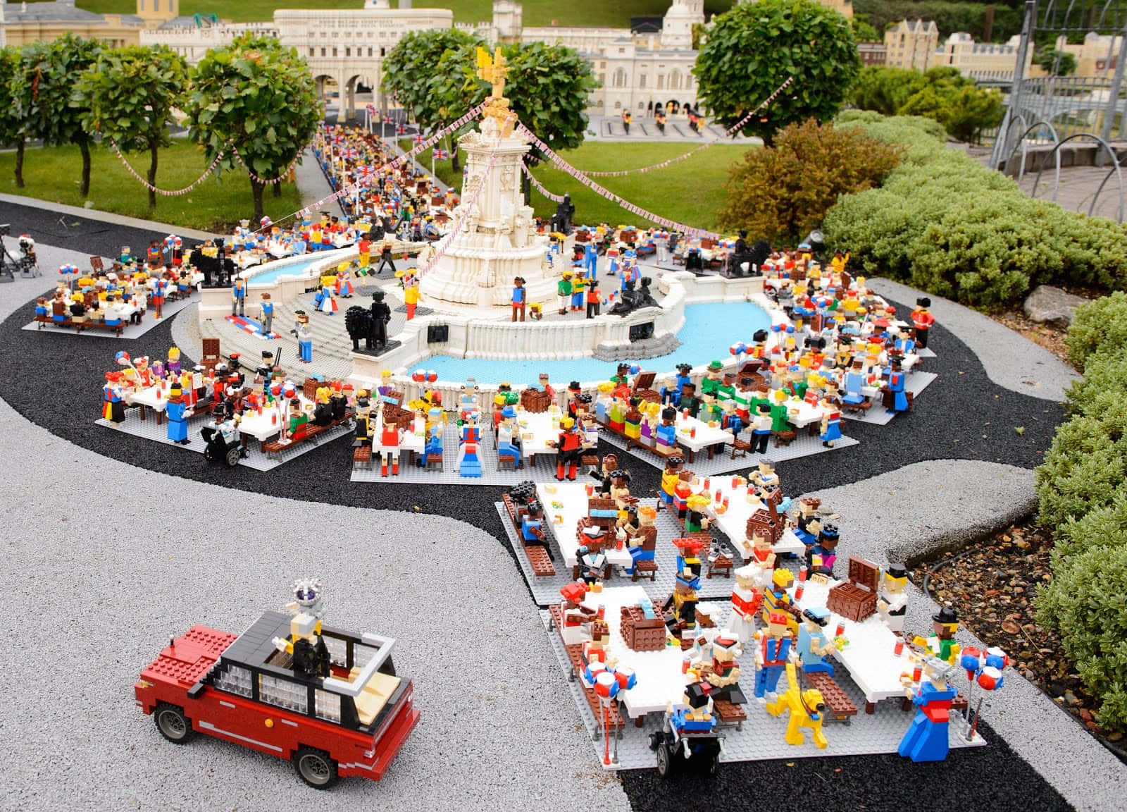 歡迎來到樂高樂園，每一天都是魔法冒險！ 壁紙 — 一個熱鬧的LEGO公園場景，小人物在中央噴泉周圍用餐，周圍環繞著綠植和一輛紅色LEGO汽車。