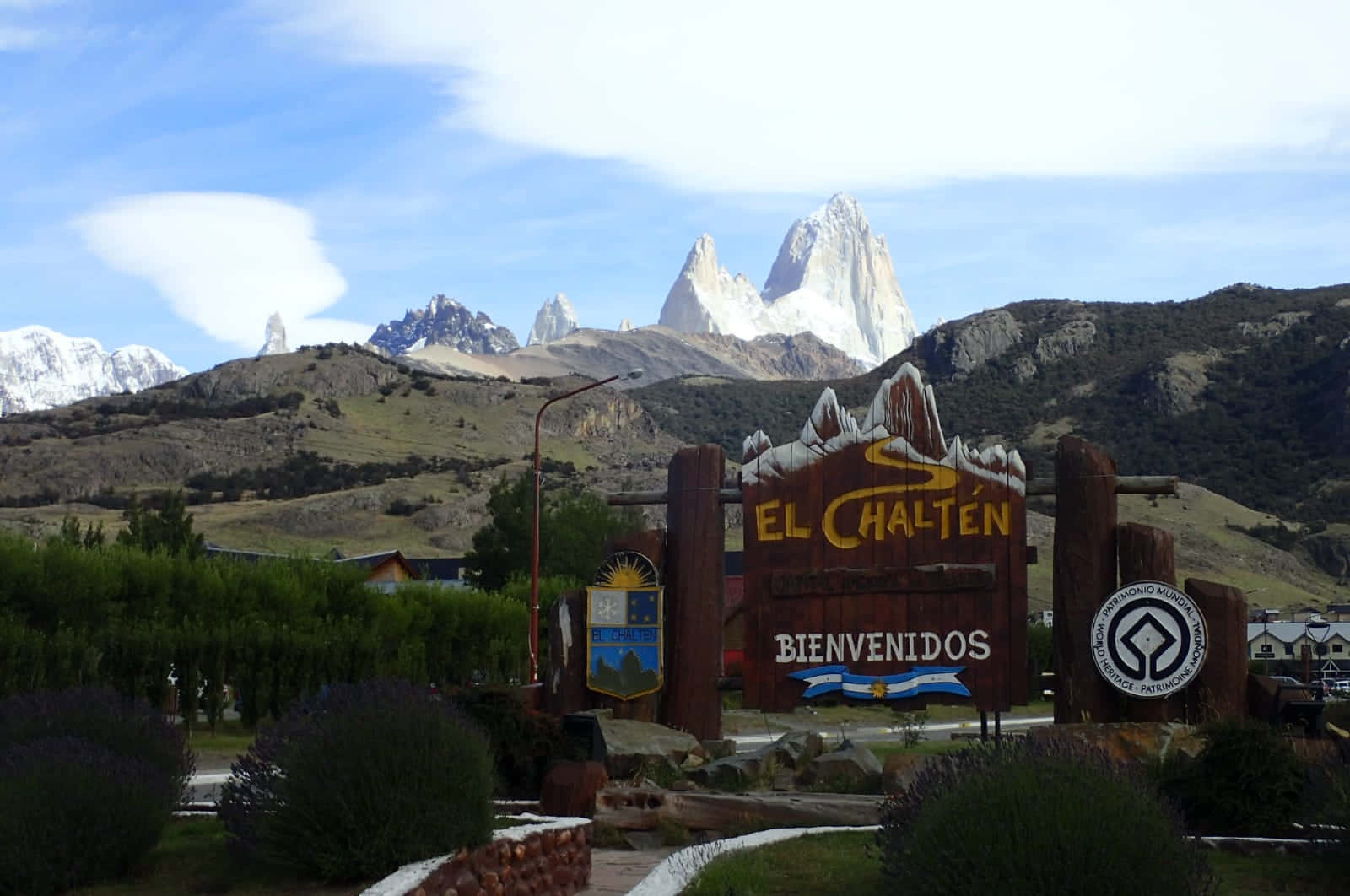 歡迎來到艾爾查滕——阿根廷的山間天堂 壁紙 — 一個木製的 El Chaltén 歡迎標誌矗立在明亮的藍天下，背景是高聳的雪山。