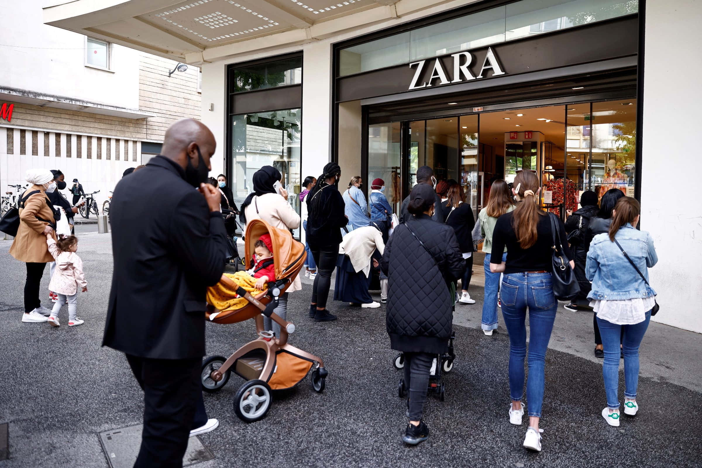 歡迎光臨 Zara！別錯過最新時尚潮流！ 壁紙 — 一群購物者喺城市行人路上，推住嬰兒車、着住休閒服裝，喺Zara舖頭外面排隊。