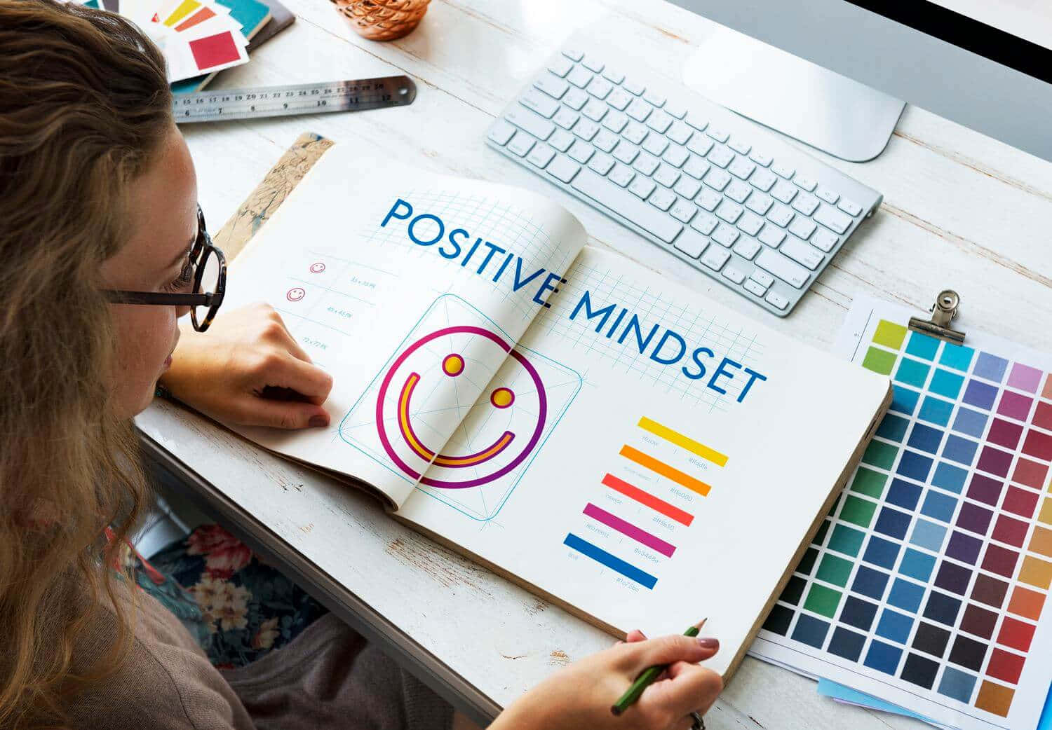 正向思考筆記微笑圖 壁紙 — 設計師在名為「Positive Mindset」的筆記本上草繪一個色彩繽紛的笑臉，周圍環繞著色板和鍵盤。