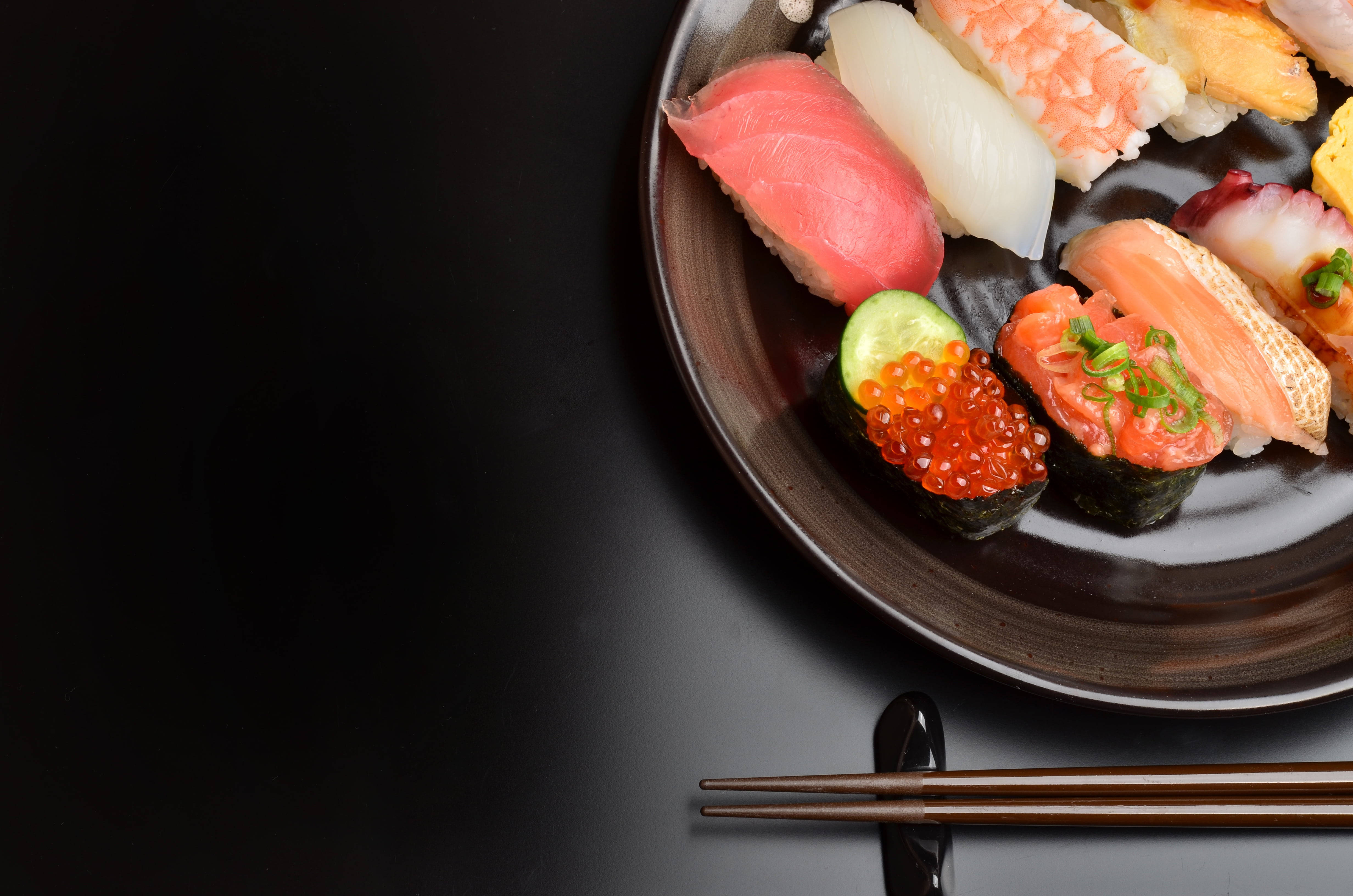 正宗日式壽司美食 壁紙 — 一個深色碟子上放著各種色彩繽紛的壽司，包括吞拿魚、三文魚和魚子，旁邊放著筷子。