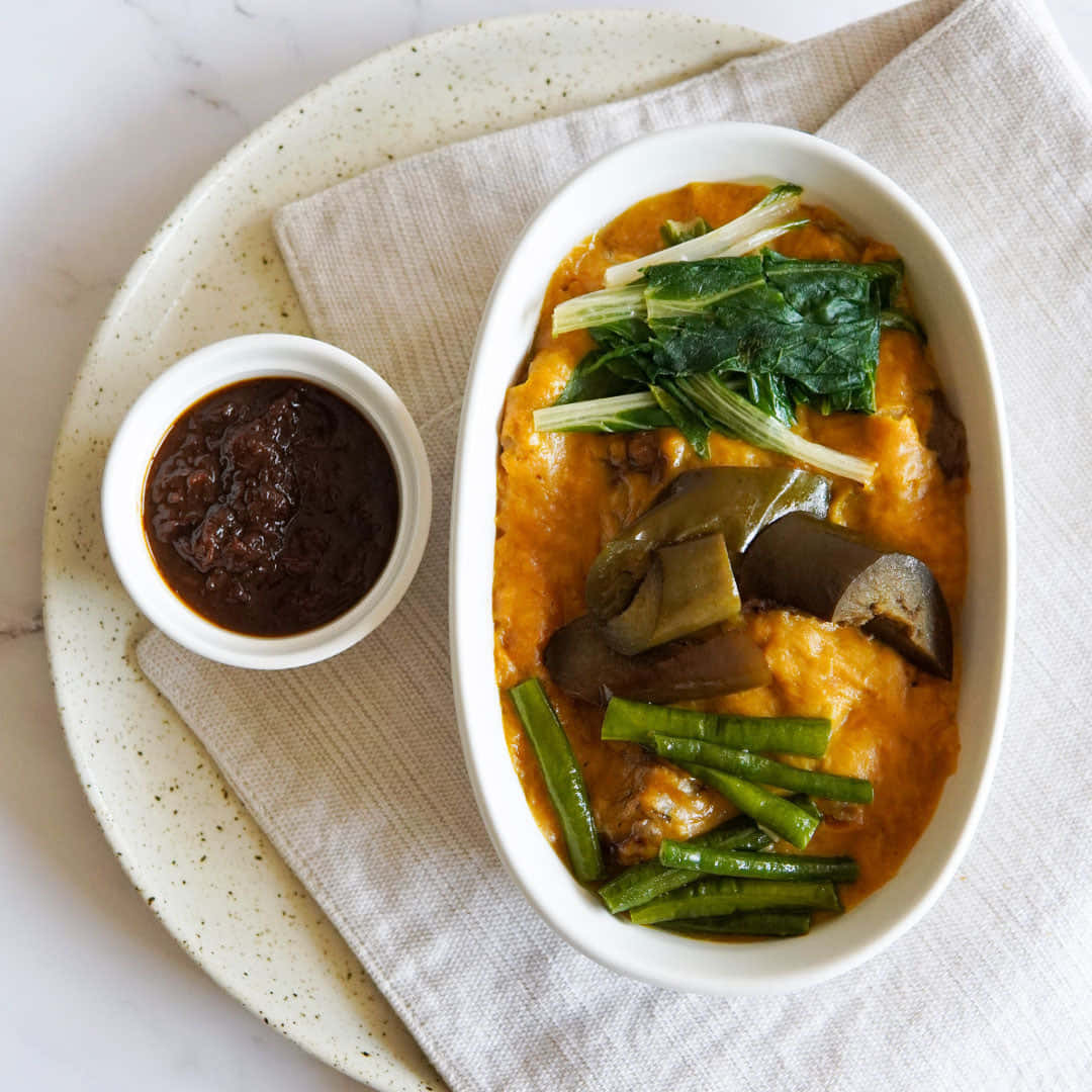 正宗菲律賓 Kare-Kare 燉菜配蝦醬 壁紙 — 一碗充滿活力的橙色咖喱，配有茄子、青豆和菠菜，盛在白色碗中，旁邊有深色醬汁，放在斑點盤上。