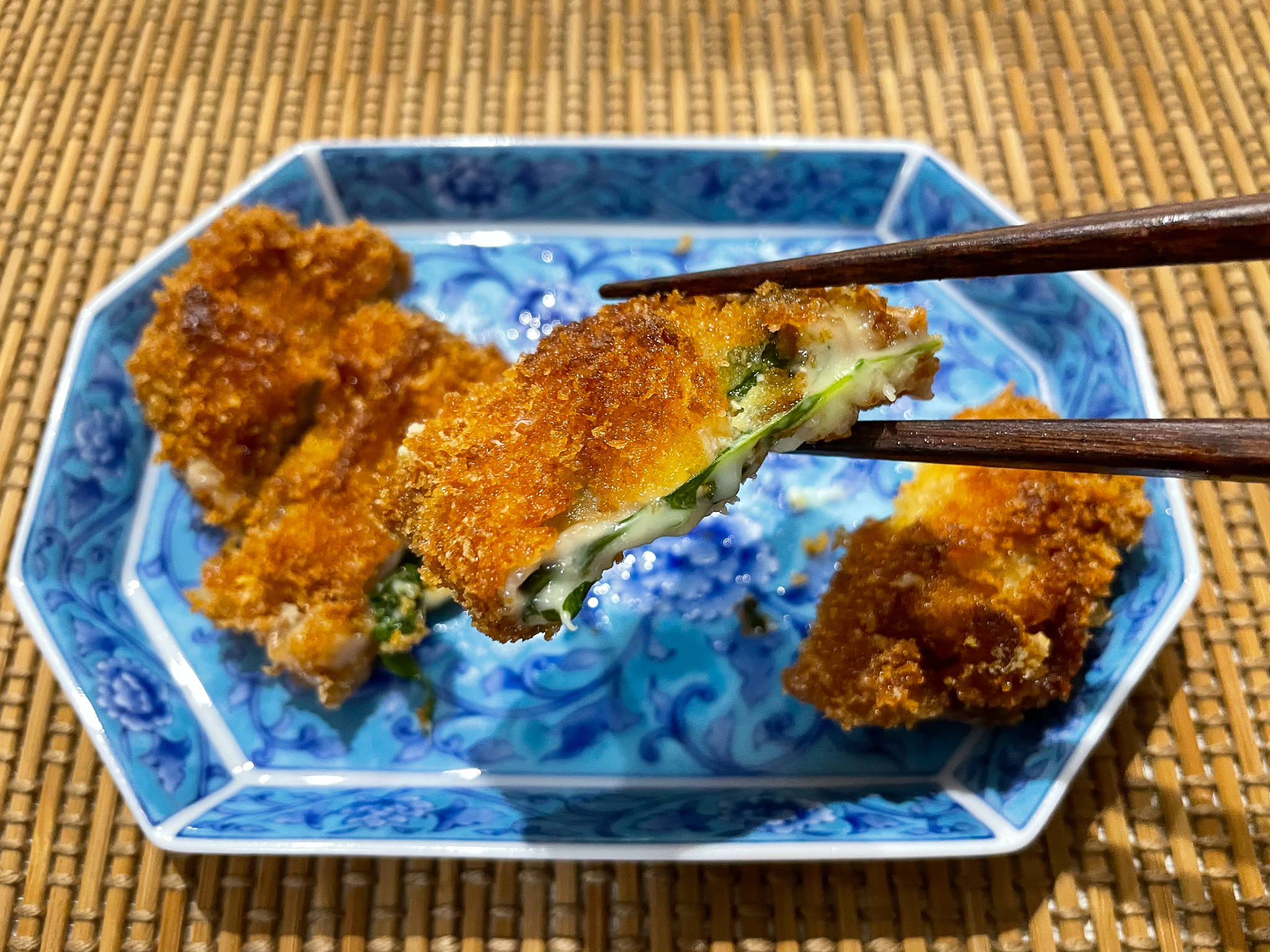 正宗酥脆豬排配奶油餡 壁紙 — 金黃色的炸雞塊，內有綠色餡料，用筷子夾起，放在竹蓆上的藍色圖案碟子上。