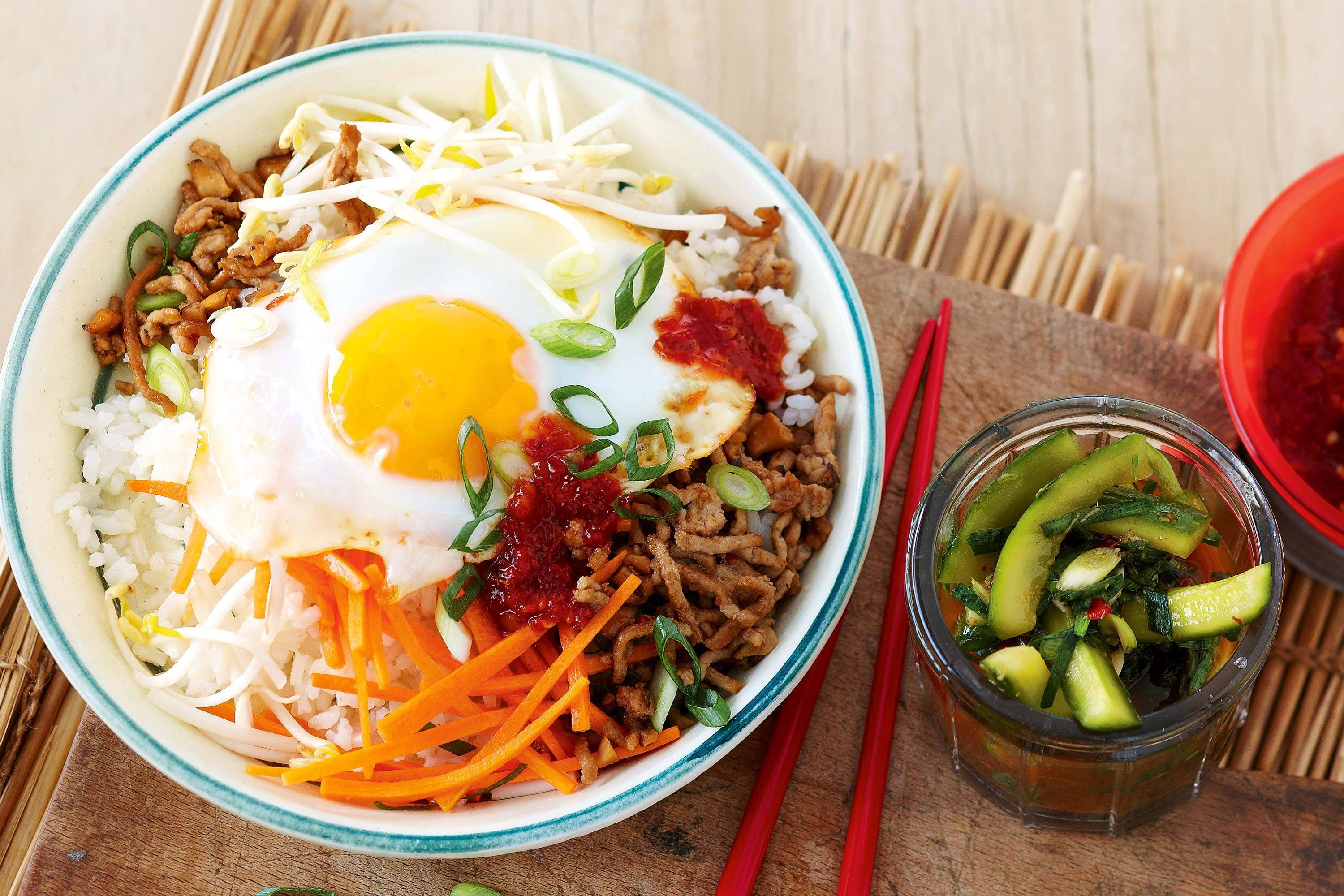 正宗韓國料理——拌飯 壁紙 — 一碗色彩繽紛的韓國石鍋拌飯，上面放有太陽蛋、豆芽、紅蘿蔔和辣醬，旁邊有醃黃瓜和紅色筷子，放在木墊上。