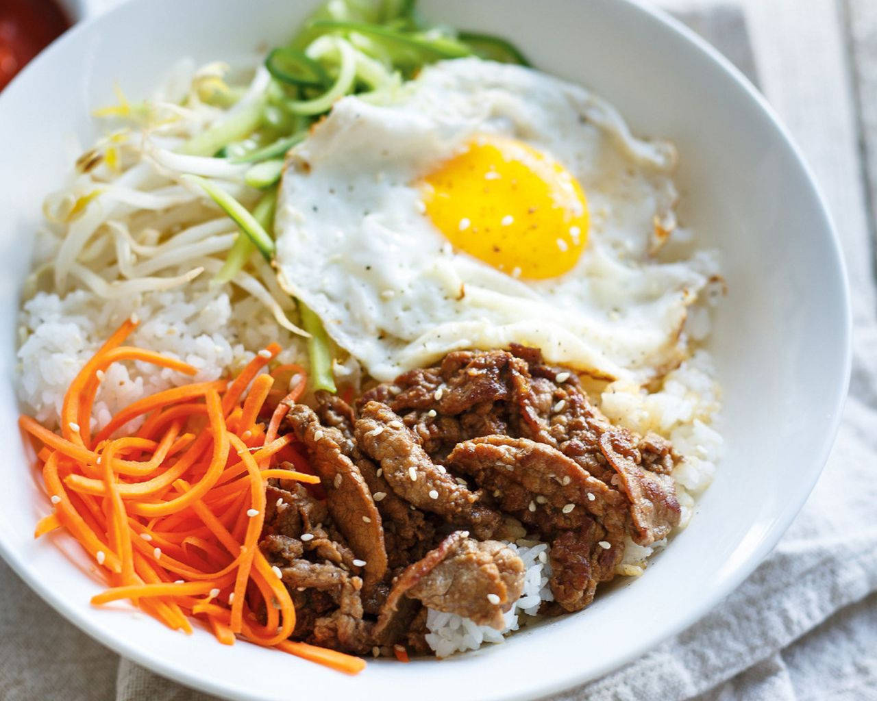 正宗韓國料理：拌飯配細條蔬菜 壁紙 — 一碗色彩繽紛的韓國石鍋拌飯，上面放有太陽蛋、豆芽、紅蘿蔔和調味牛肉，搭配白飯。