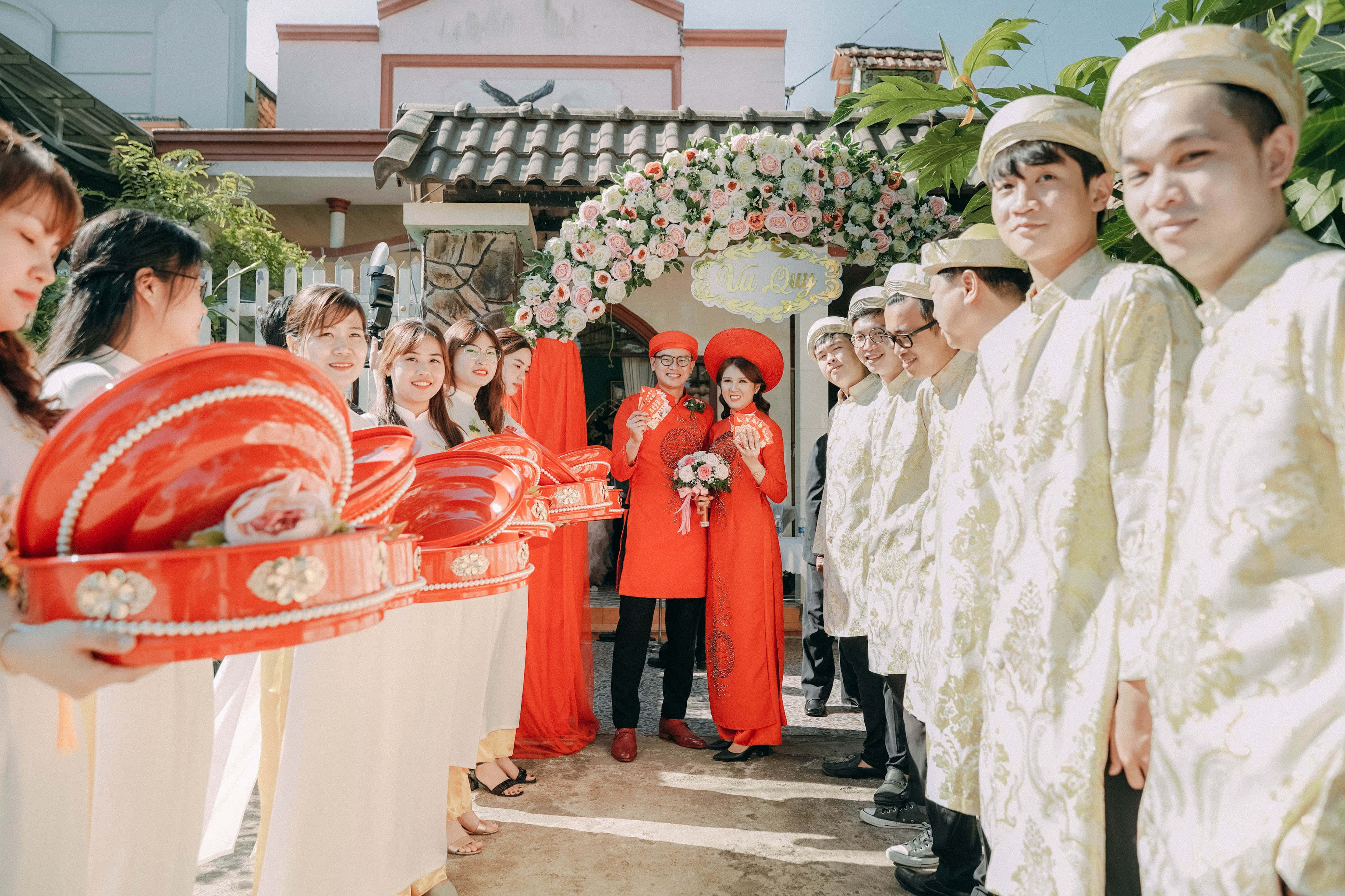 正式婚宴上的優雅伴郎團 壁紙 — 越南婚禮遊行中，新娘身穿紅色奧黛，新郎身穿白色奧黛，在花拱下，伴隨者手持紅色儀式托盤。
