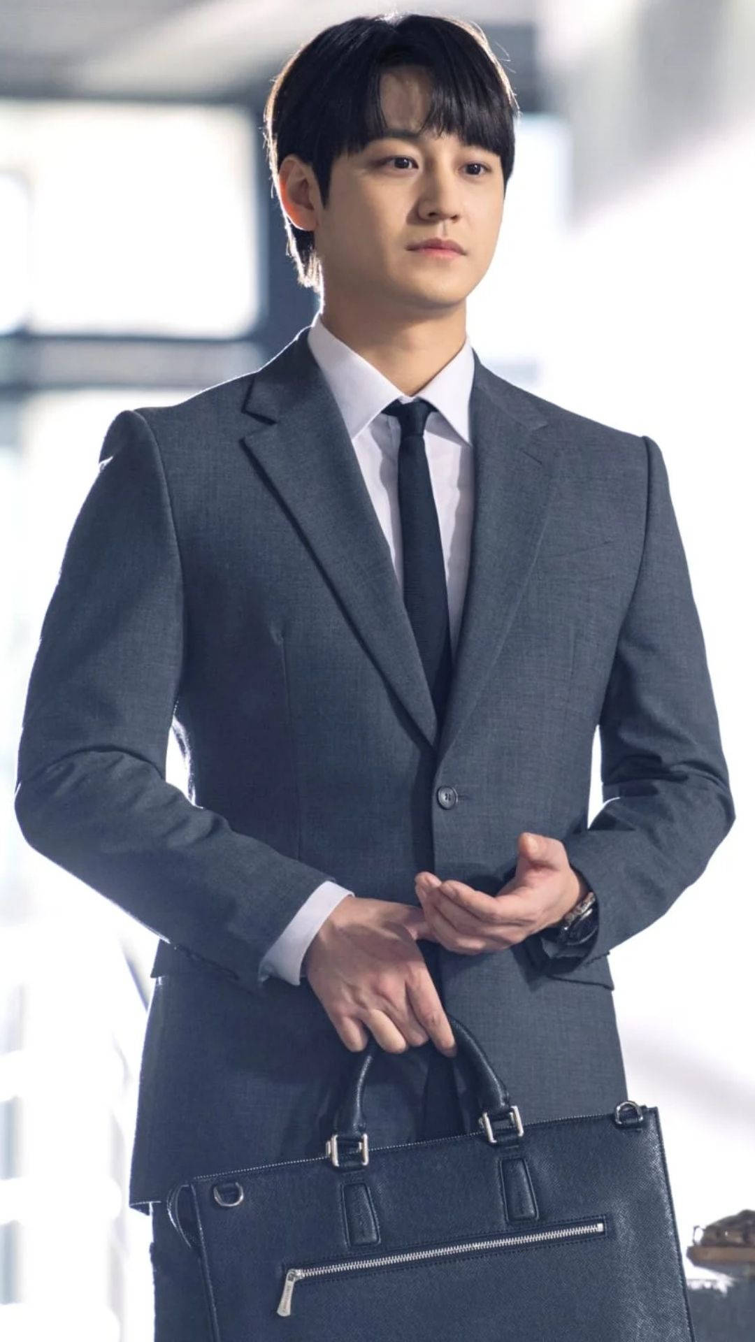 正式著裝的金範 壁紙 — 一位穿著灰色西裝、打扮俐落的男士，在明亮模糊的背景前，表情嚴肅地拿著一個黑色公事包。
