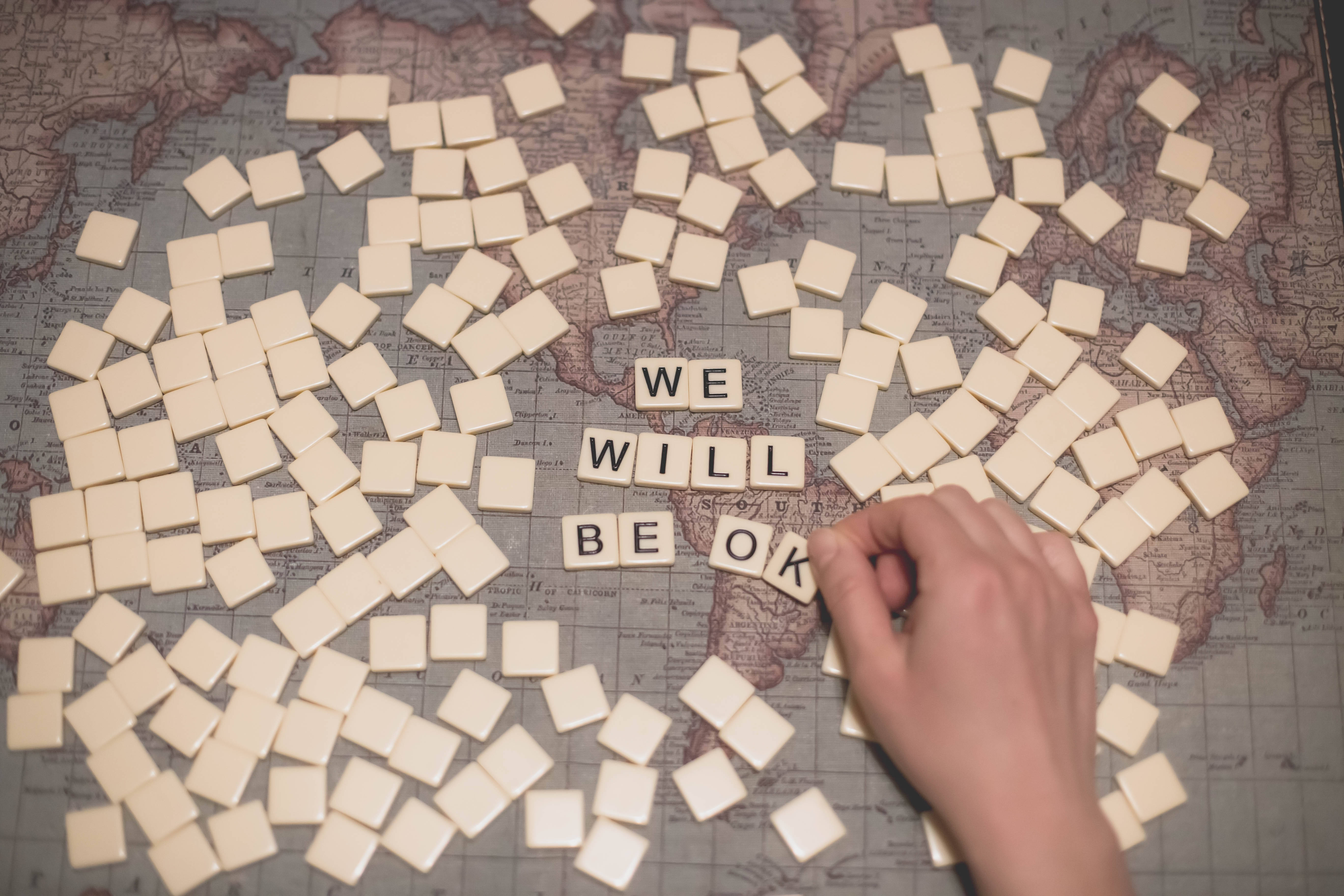 正面心態 壁紙 — 一隻手將 Scrabble 字母牌放在復古世界地圖上，拼出 'we will be ok'，周圍散落著字母牌。