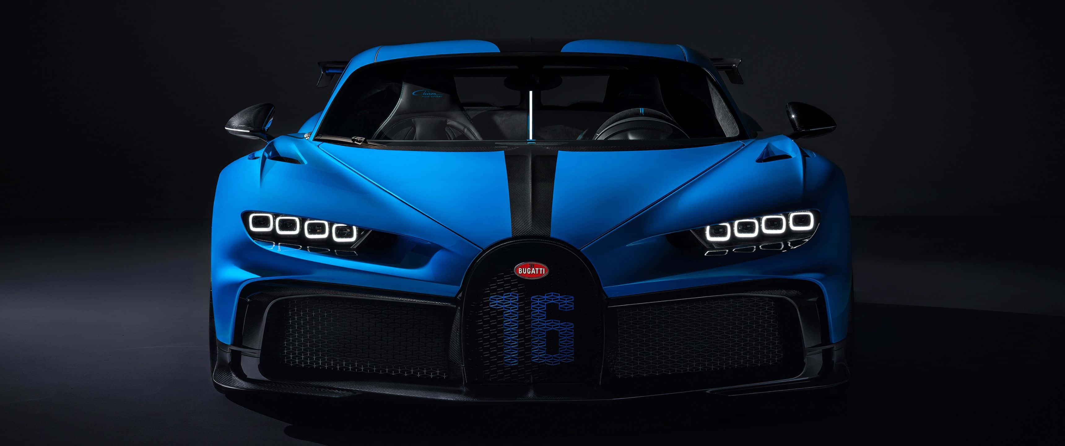 步入奢華，盡享這款 3440x1440p 宾利背景 壁紙 — 一輛引人注目的藍色Bugatti Chiron Super Sport 300+，前格柵上有16號標誌，在黑暗的攝影棚背景下被銳利的車頭燈照亮。