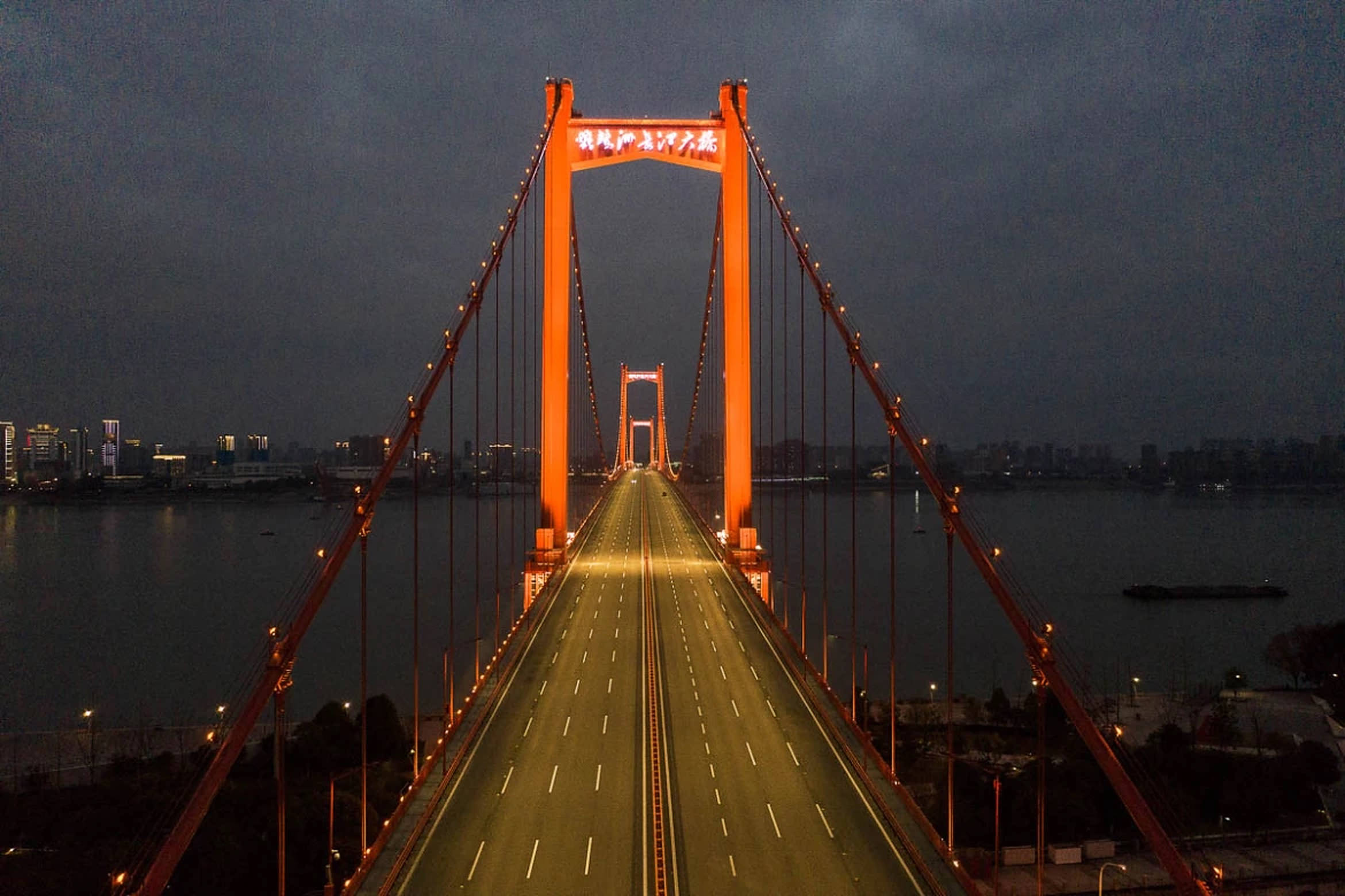 武漢 Yingwuzhou 長江大橋 壁紙 — 一座高聳的橙色吊橋在暮色中發光，空蕩的車道延伸向遠方的城市天際線。
