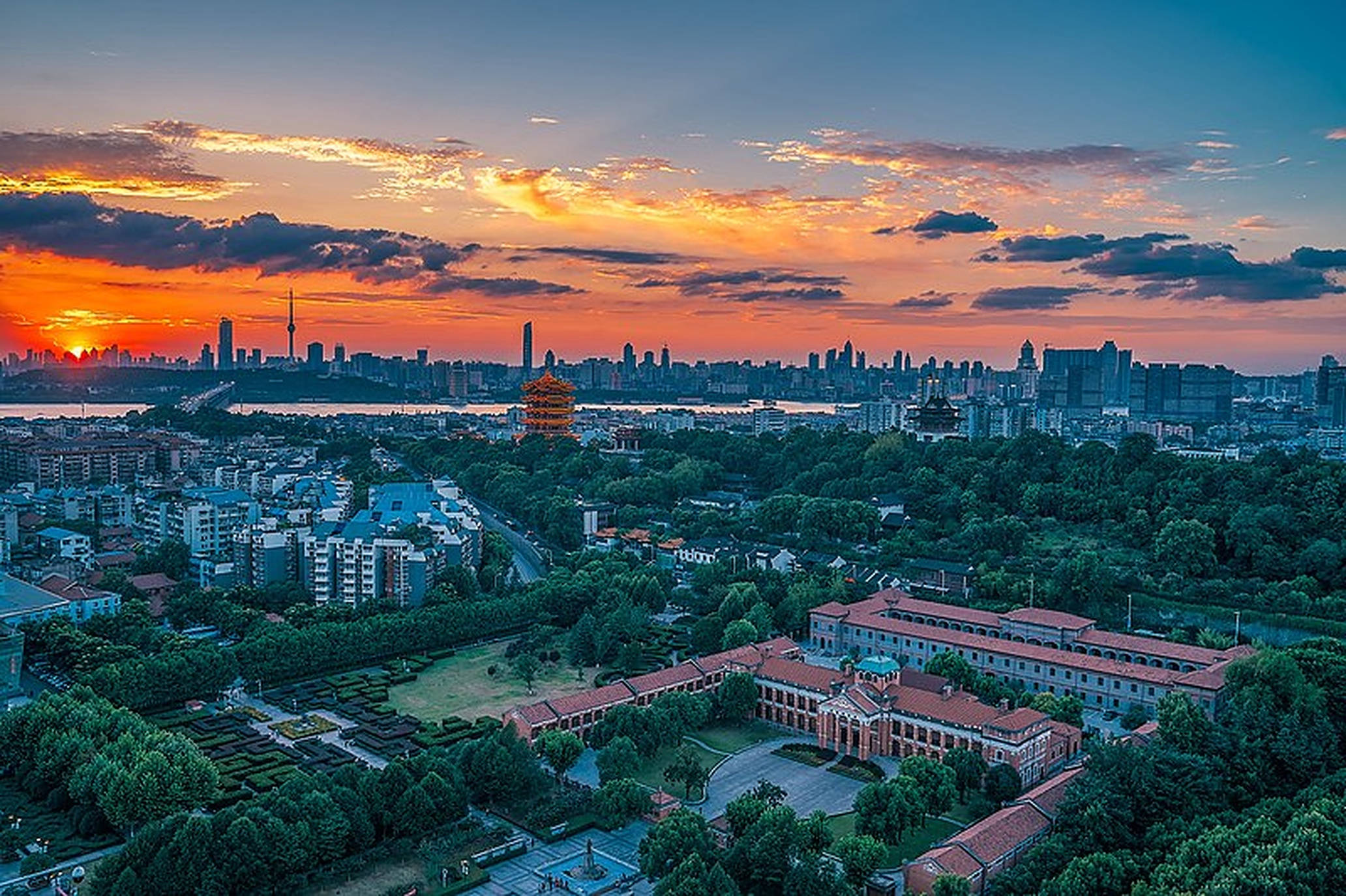 武漢城市橙色夕陽 壁紙 — 從高空俯瞰日落時分的城市景觀，河流上方有橙色雲彩，歷史建築被茂密的綠樹環繞。