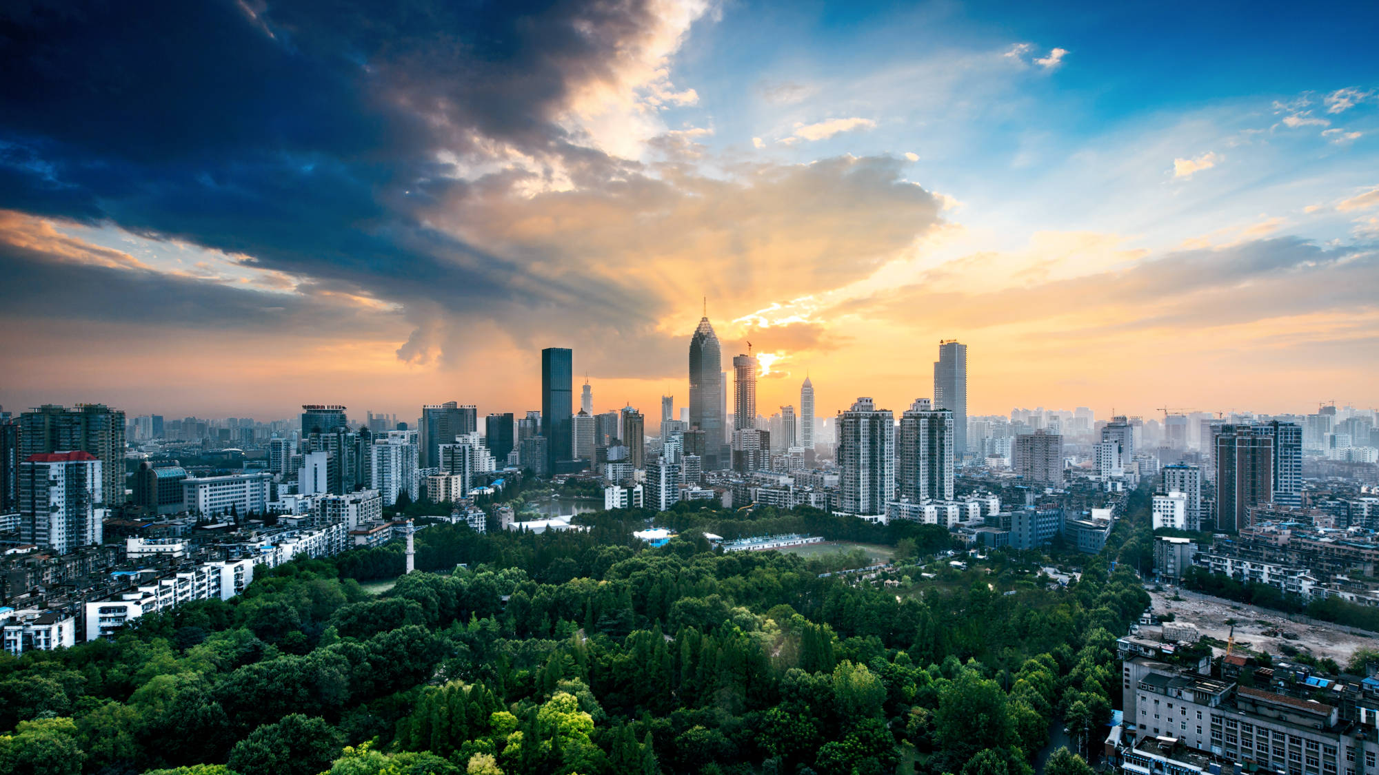 武漢城市風景 壁紙 — 夕陽下充滿活力的城市天際線，金色光芒穿透戲劇性的雲層，照耀在鬱鬱蔥蔥的綠色公園之上。