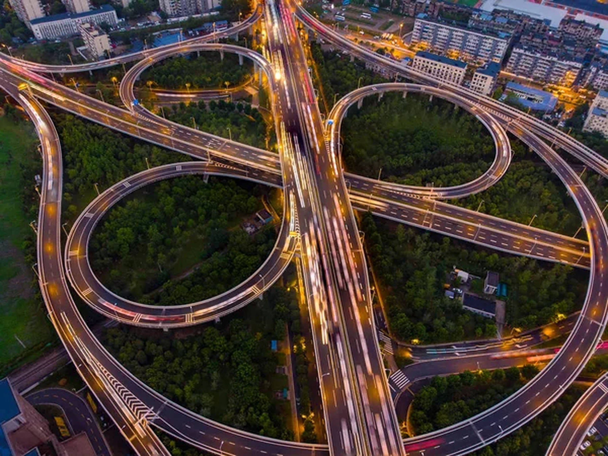 武漢環形高架路 壁紙 — 黃昏時分複雜高速公路交匯處的鳥瞰圖，移動中的汽車發出閃爍的光軌，蜿蜒穿過茂密的綠植。