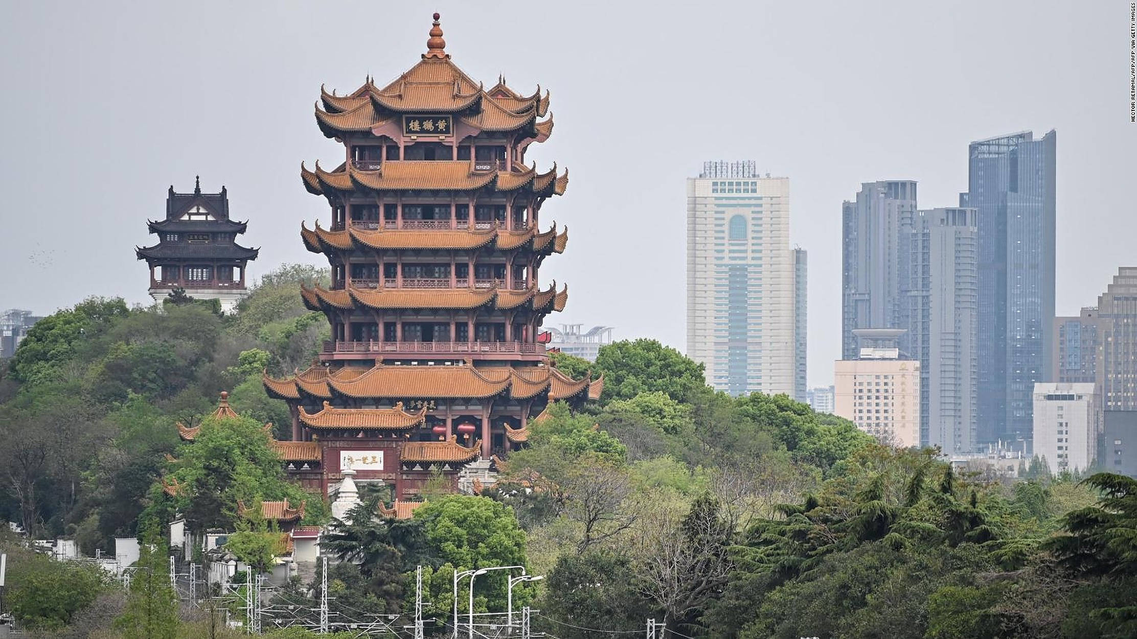 武漢黃鶴樓 壁紙 — 一座擁有橙色屋頂的傳統中式寶塔矗立在鬱鬱蔥蔥的綠色山丘上，被現代摩天大樓環繞，天空多雲。