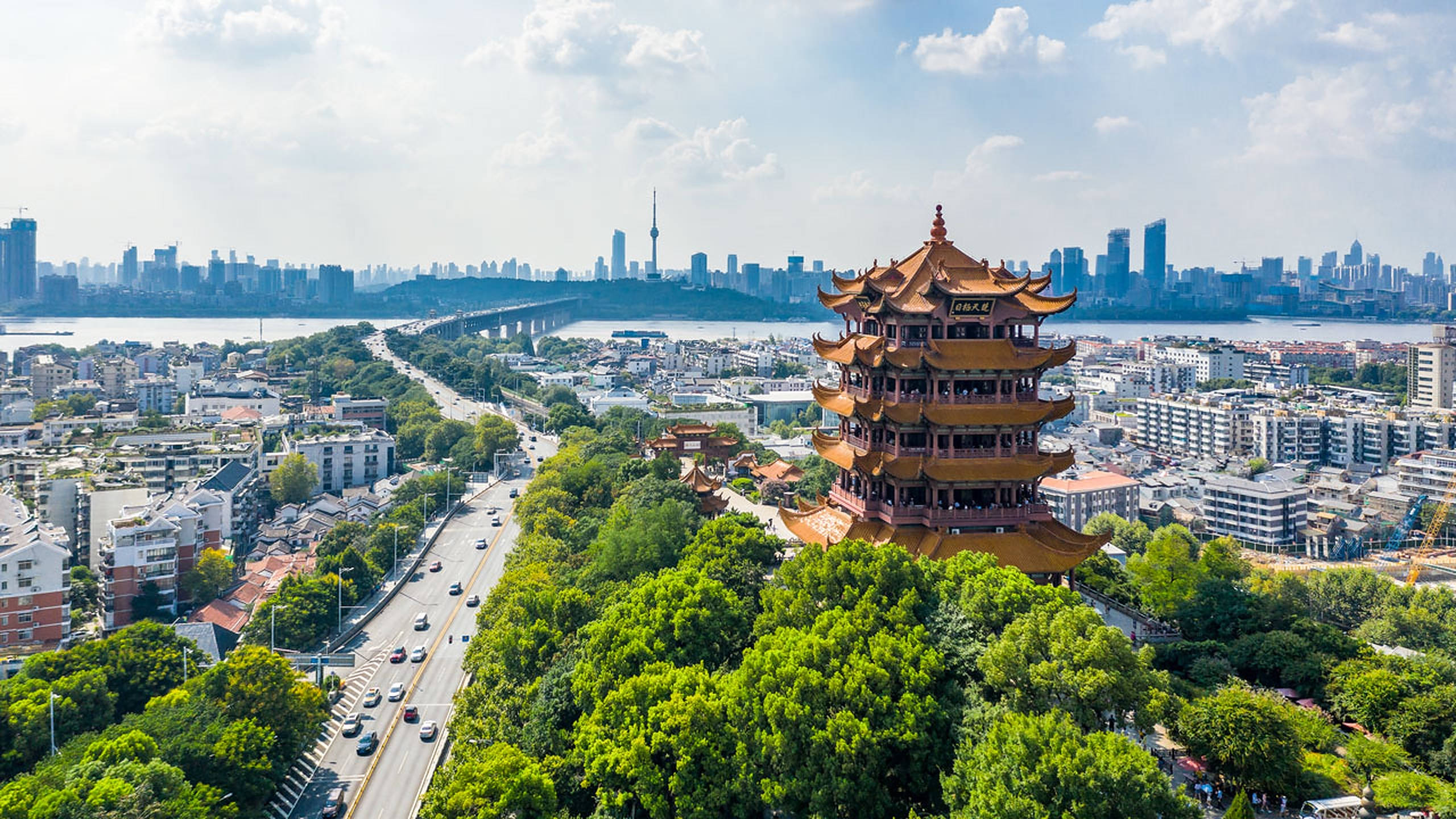 武漢黃鶴樓日間壯觀景觀 壁紙 — 喺明亮藍天下，繁華現代城市景觀中，被茂密綠樹環繞嘅傳統中國寶塔嘅航拍視圖。