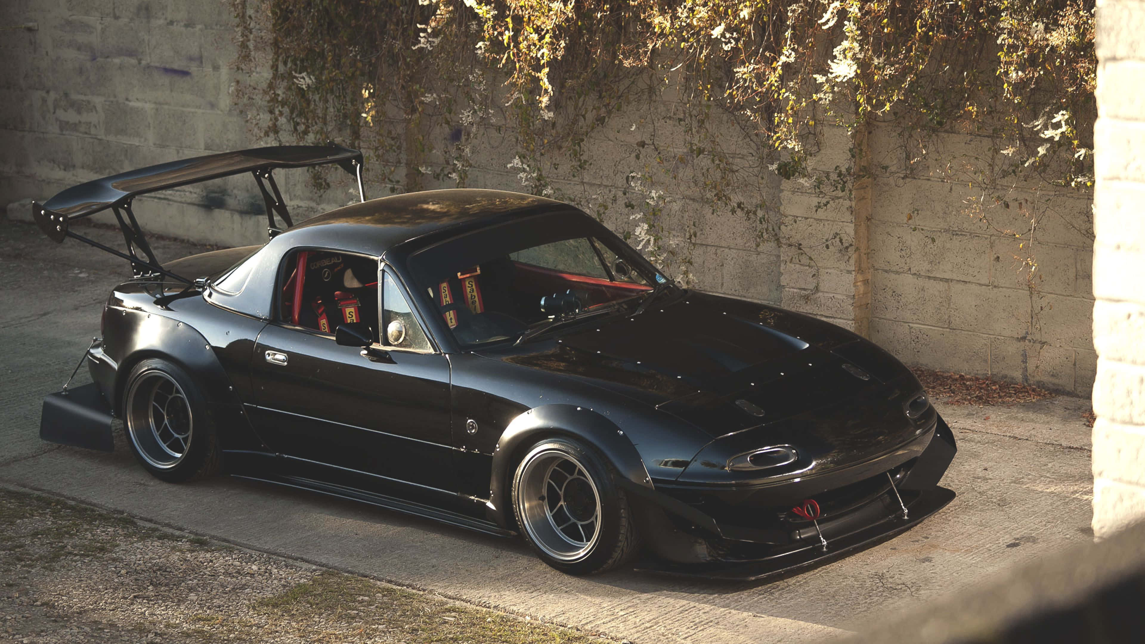 歷久彌新的經典敞篷跑車：馬自達MX-5 Miata 壁紙 — 一架時尚黑色改裝跑車，帶有大型後擾流板，停泊在藤蔓覆蓋的混凝土牆下。