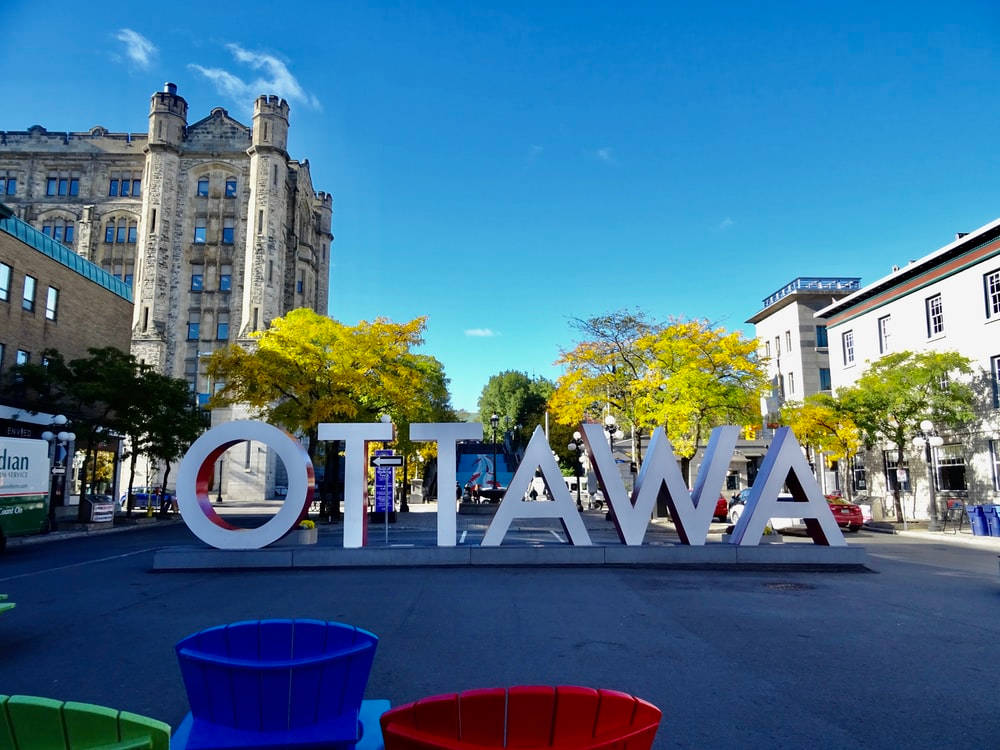 歷史性拜沃德市場中的鮮豔渥太華標誌 壁紙 — 大型白色「OTTAWA」字母矗立在陽光明媚的城市廣場上，周圍有秋樹和歷史建築，背景是清澈的藍天。