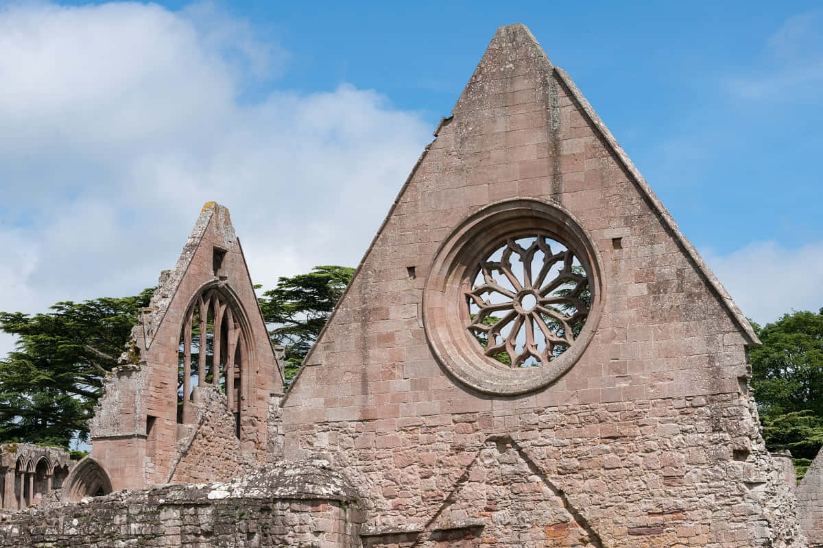 Gothic Architecture 桌布 壁紙 — 歷史悠久的教堂的陽光普照的石頭廢墟，帶有突出的玫瑰窗和尖拱，襯著藍天。