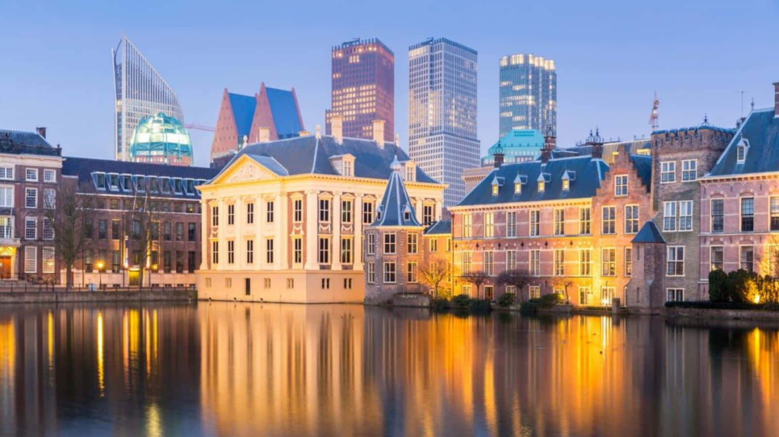 Netherlands 桌布 壁紙 — 歷史悠久的運河房屋在暮色中映照著溫暖燈光，倒映在平靜的水面上，背景是現代城市的天際線。