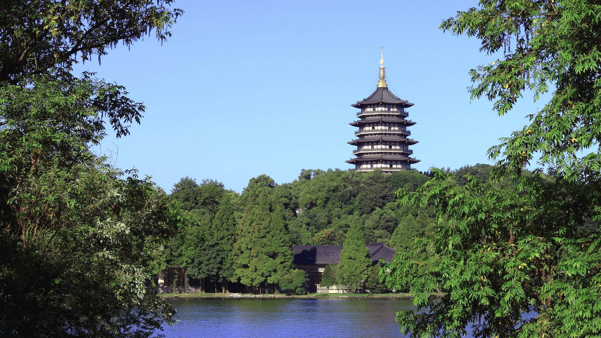 歷史杭州雷峰塔 壁紙 — 一座傳統多層寶塔聳立在翠綠樹木之上，旁邊是平靜的藍色湖泊，天空晴朗。