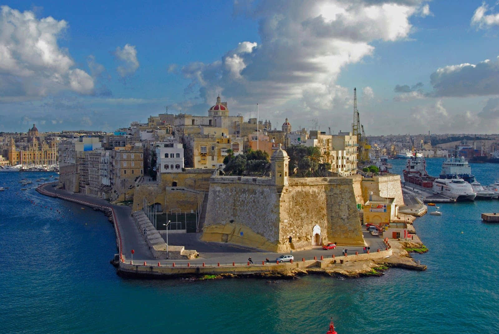 Malta 桌布 壁紙 — 歷史石堡壘和沿海城市景觀的鳥瞰圖，在部分多雲的天空下，有碧綠的海水和停泊的船隻。
