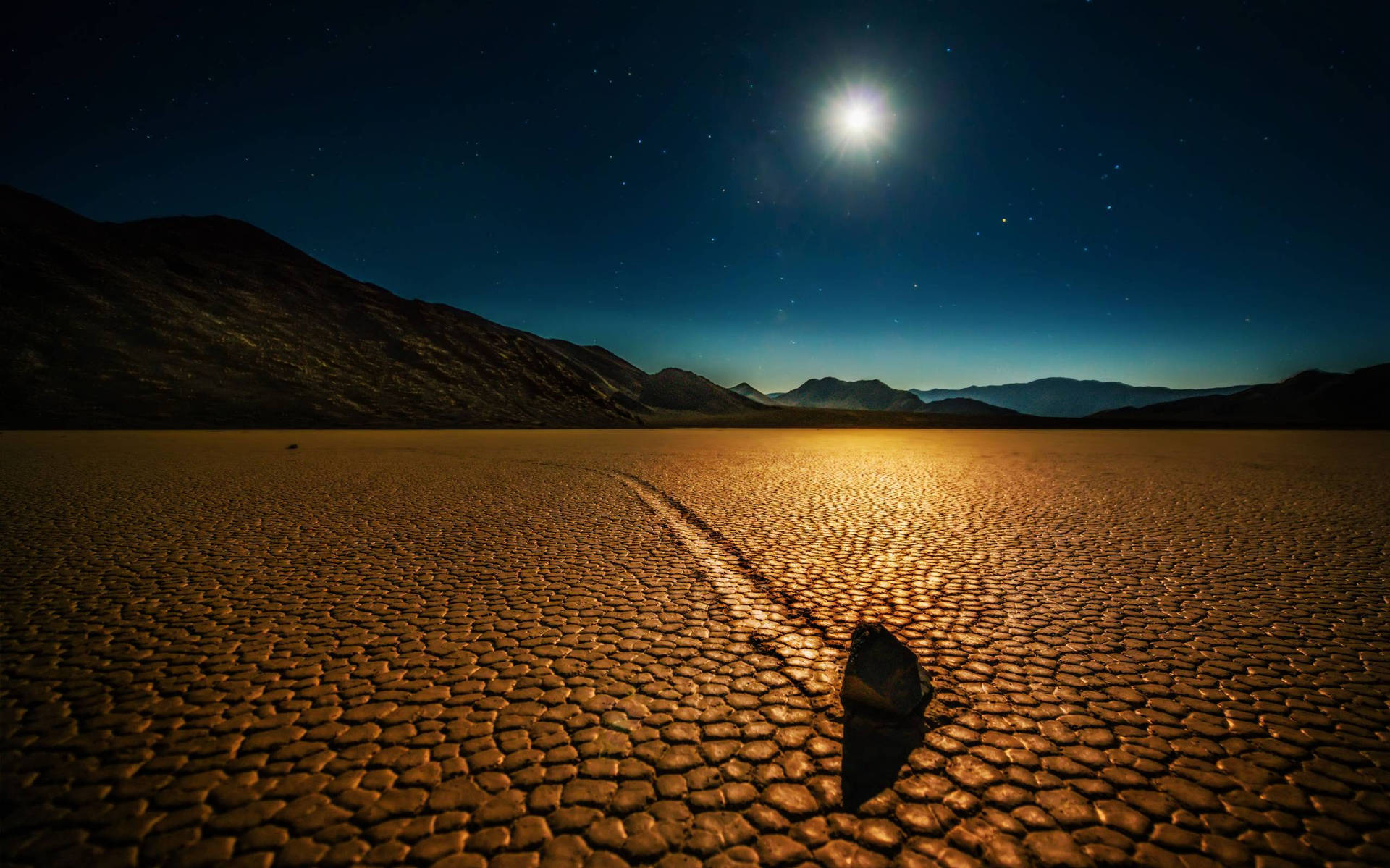 死亡谷賽車平地月夜 壁紙 — 在明亮的月亮和繁星點點的夜空下，一塊孤獨的岩石在龜裂的沙漠地面上投下長長的陰影。