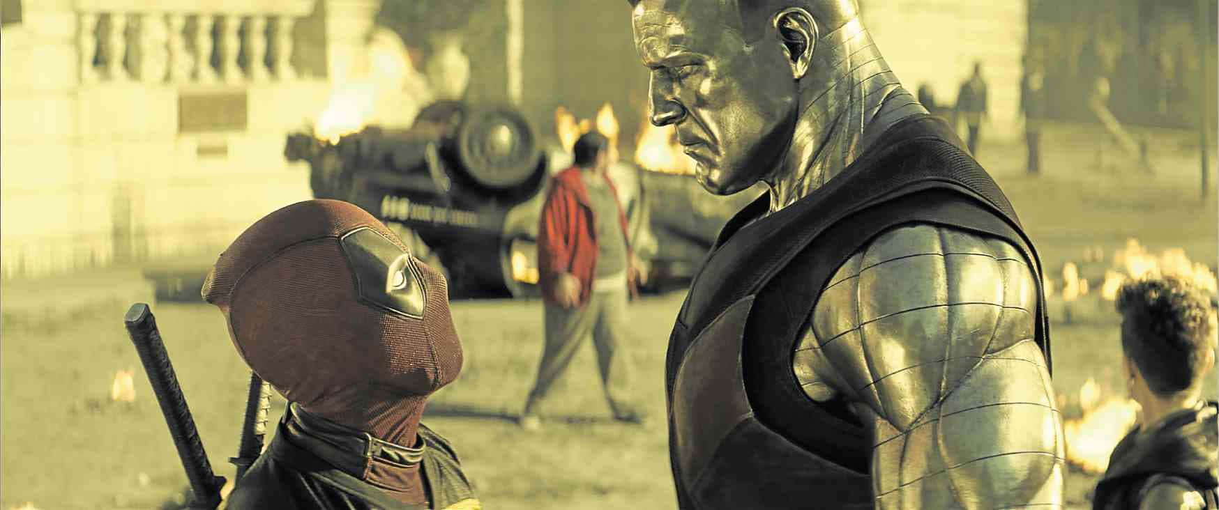 Deadpool And Colossus 桌布 壁紙 — 死侍在煙霧彌漫的戰場上與金屬裝甲反派對峙，背景有燃燒的車輛和遠處的人影。