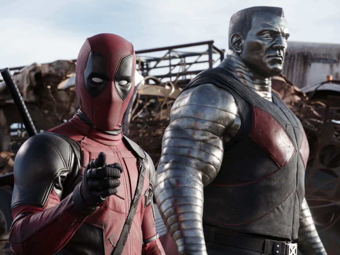 Deadpool And Colossus 桌布 壁紙 — 死侍身穿紅黑戰衣，在廢車場中指向前方，旁邊有一座金屬銀色的鋼人雕像。