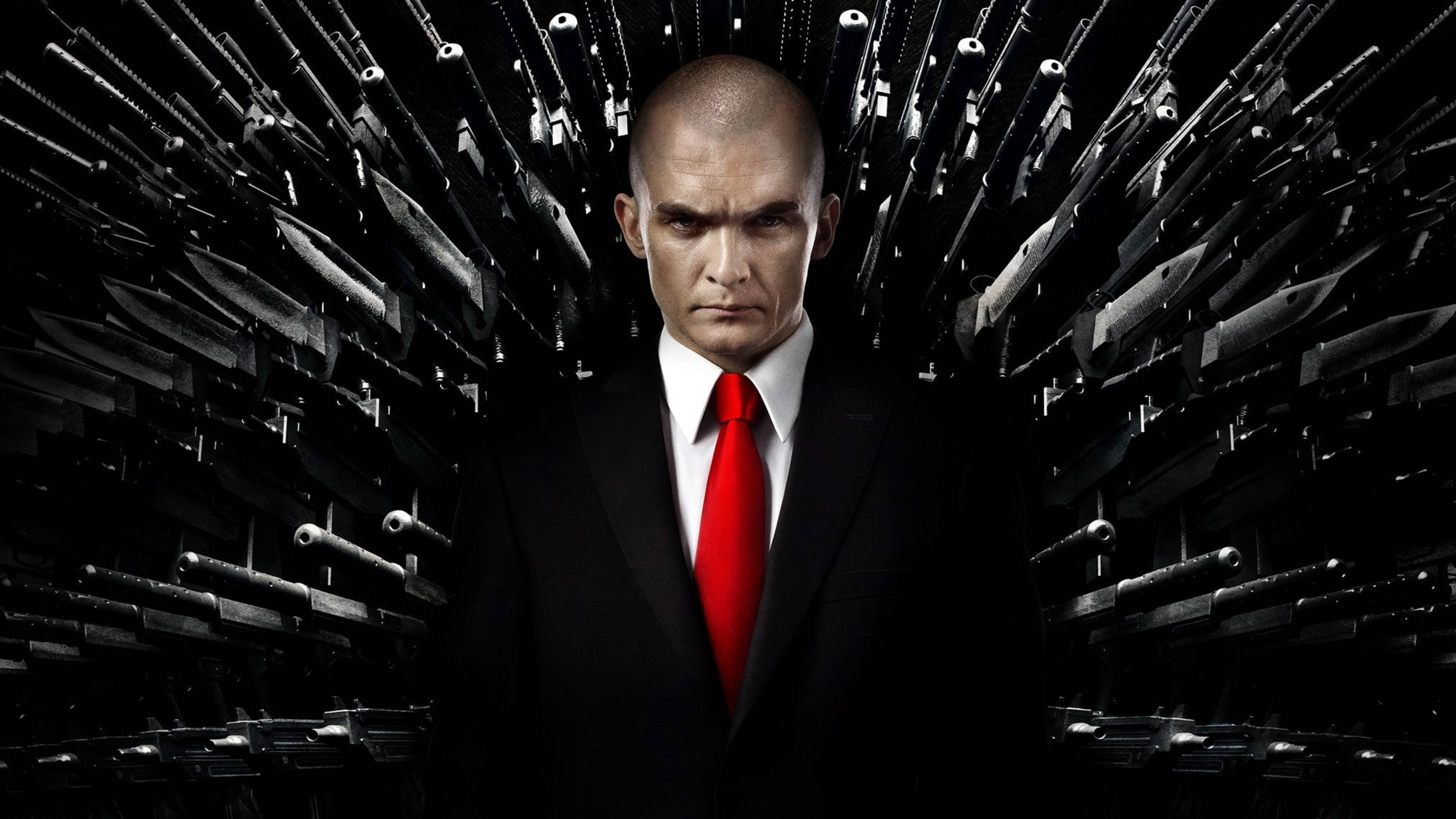 殺手47 全4K 刀槍交錯 壁紙 — 一個光頭男子身穿黑色西裝和紅色領帶，在黑暗背景前對稱排列的威脅性槍械中央站立。