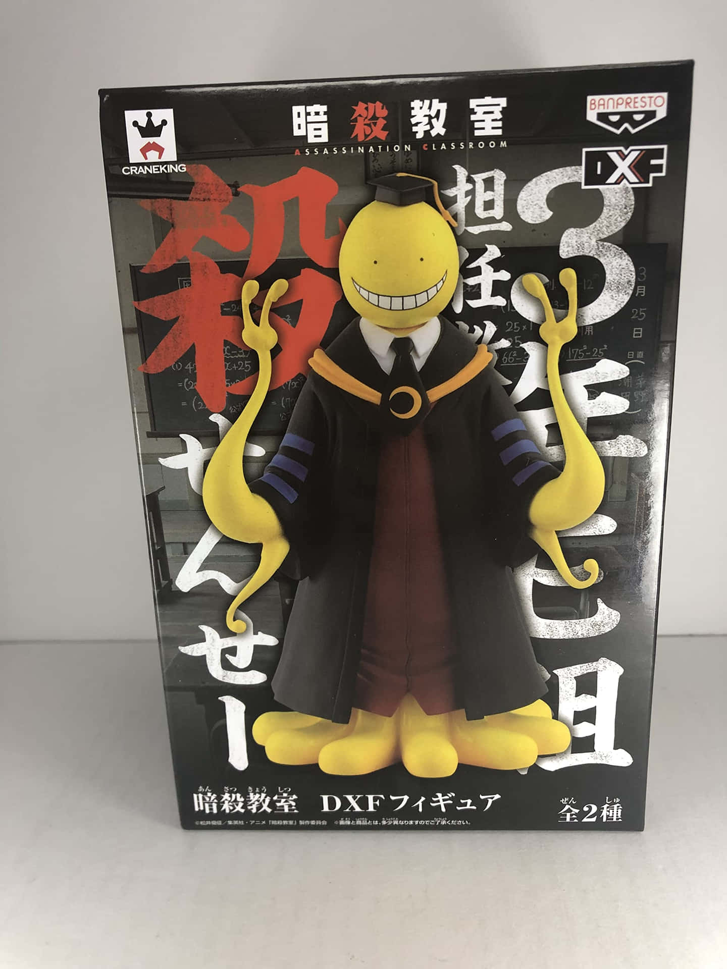 《殺老師》玩具圖 壁紙 — 一個黃色動漫人物，咧嘴大笑，手臂長長，站在黑色包裝前，包裝上有日文和紅色漢字。