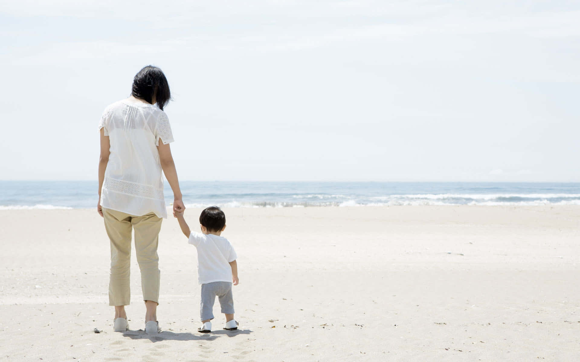 母親和幼兒在陽光明媚的海灘上手牽手走向大海。