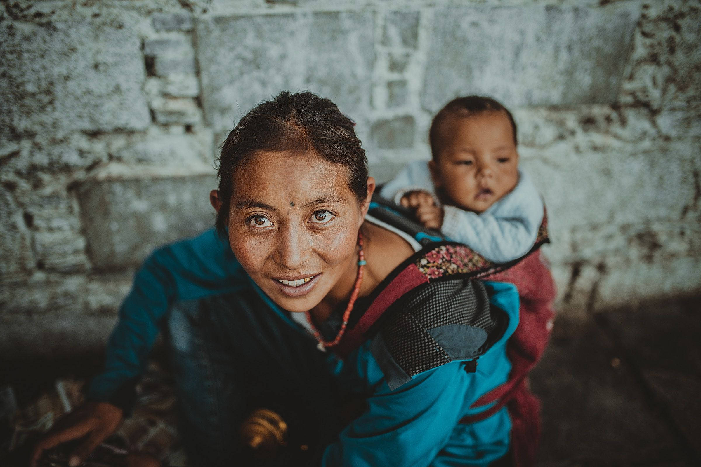 母子背負前行 壁紙 — 一位微笑的女士背著嬰兒，在風化的石牆前直視鏡頭。