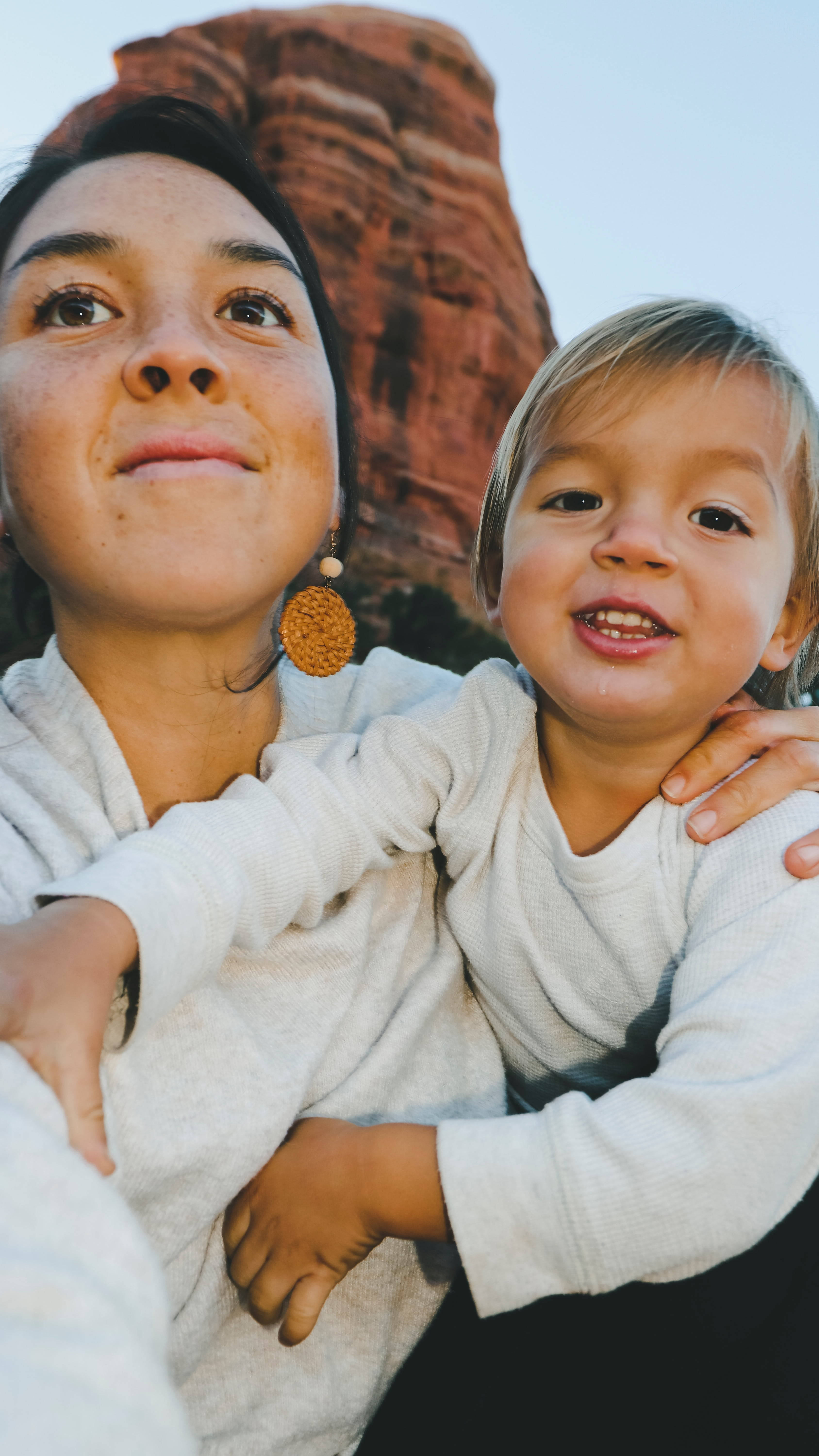 母子身穿白色 壁紙 — 一位母親和幼兒在晴朗的天空下，以紅色岩層為背景，溫暖地微笑擁抱。