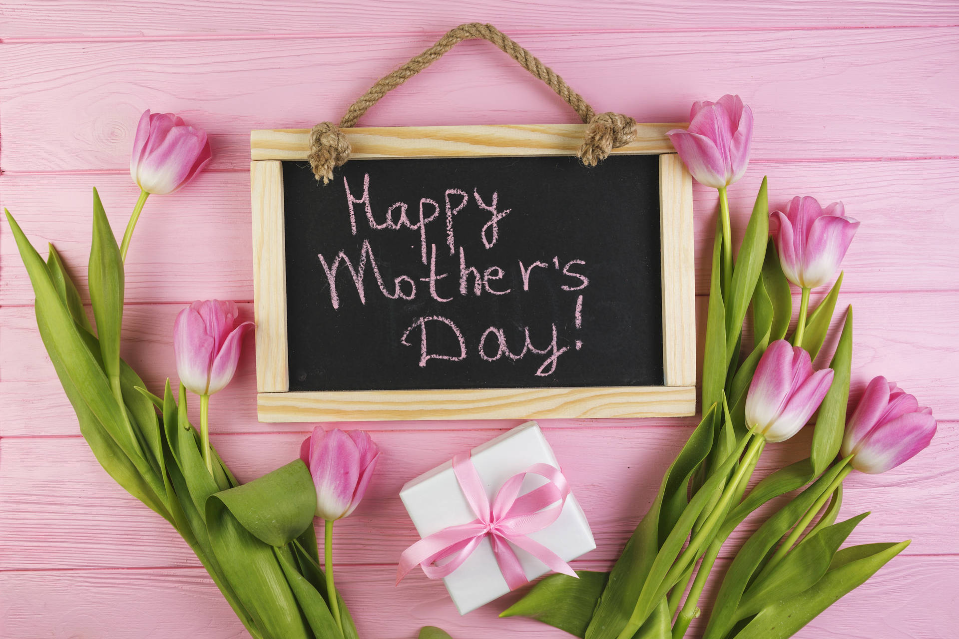 粉紅色鬱金香和禮物盒框住一塊寫有「Happy Mother's Day!」的黑板，放在粉紅色木頭上。