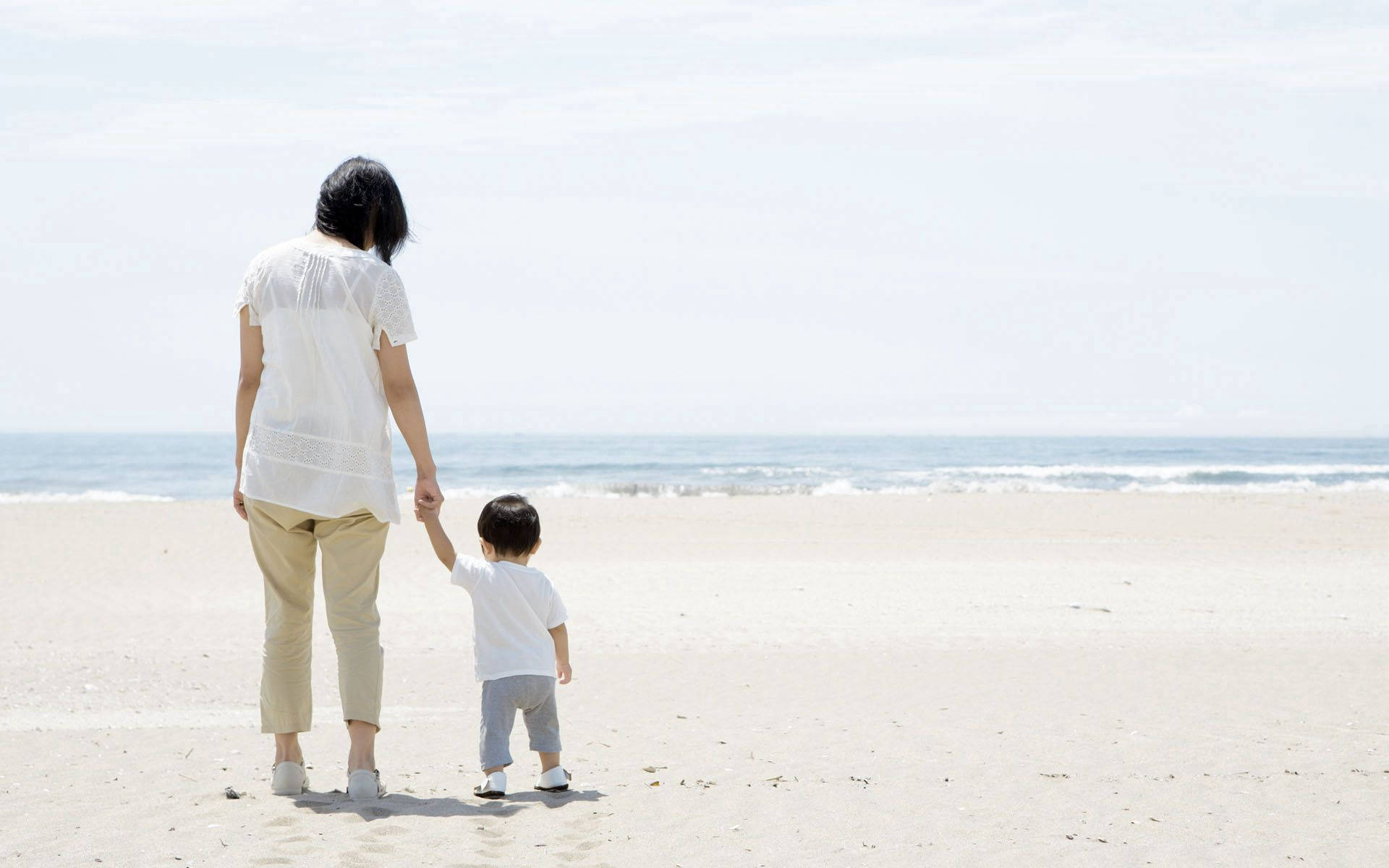 母親和幼兒手牽手在沙灘上走向海洋。