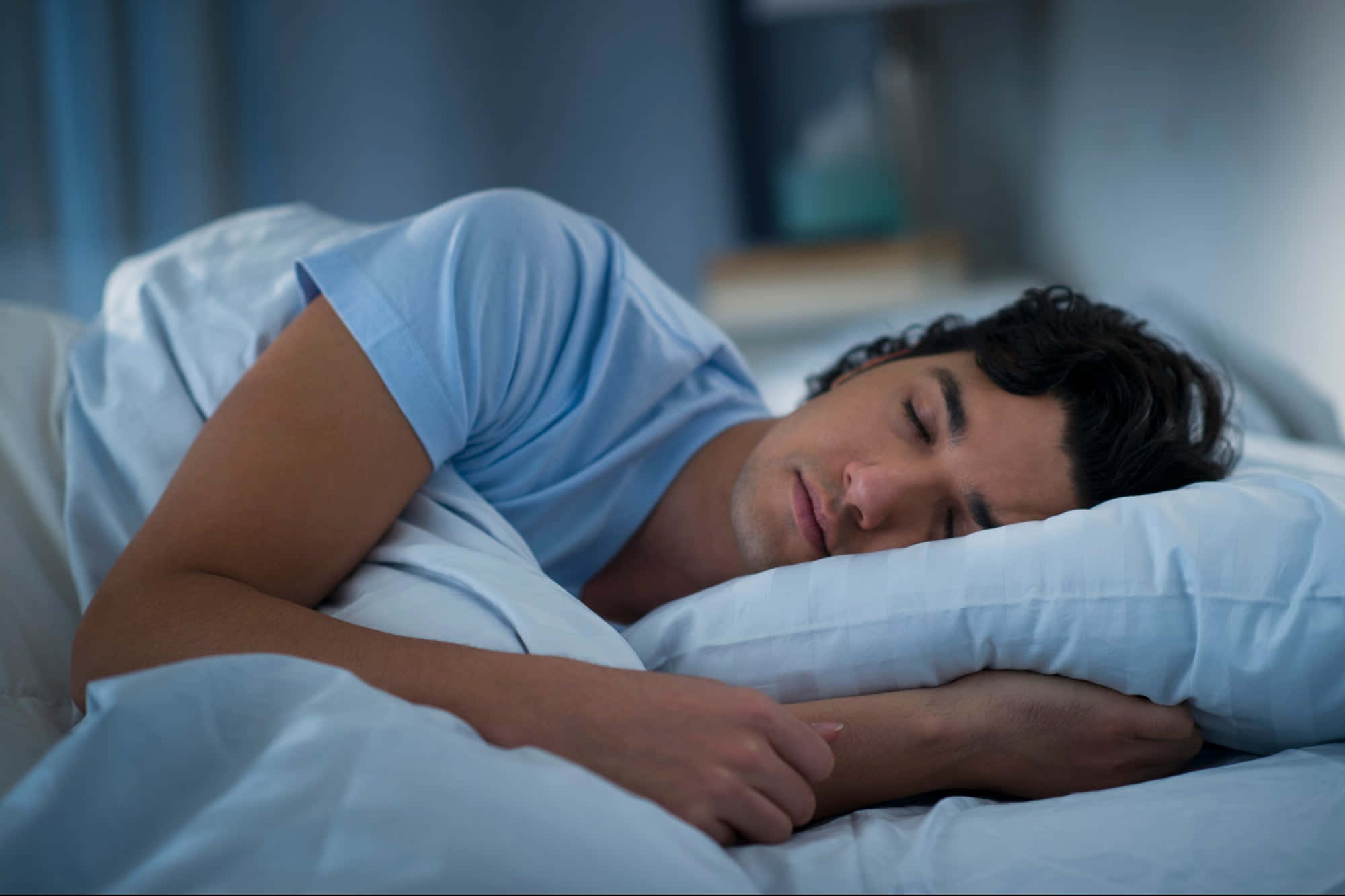 每晚獲得滿意的睡眠有助於維持身心健康 壁紙 — 一名男子在柔和燈光、藍色調和白色床品的睡房中側臥安睡。