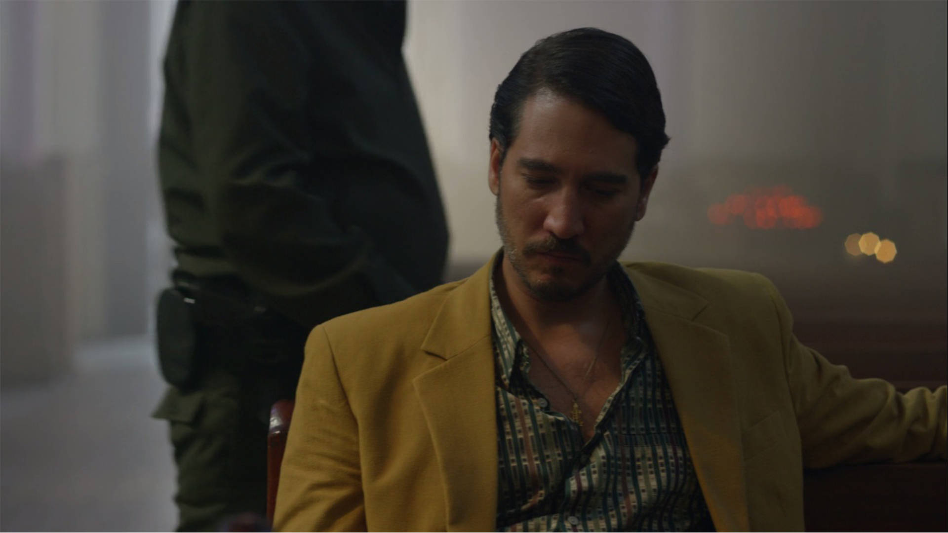 一名穿著黃色西裝外套的男子閉著眼睛坐著，一名穿制服的警員站在他身後。