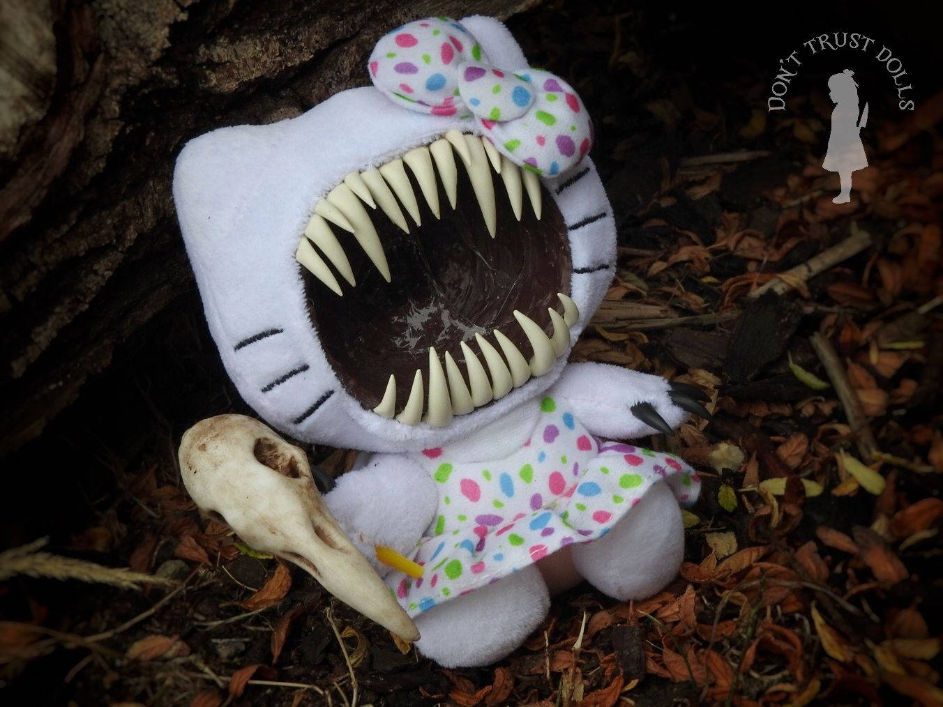 毒液Hello Kitty萬聖節 壁紙 — 一個白色毛絨玩具，類似Hello Kitty，有尖銳的牙齒，旁邊有一個骷髏頭，躺在落葉中。