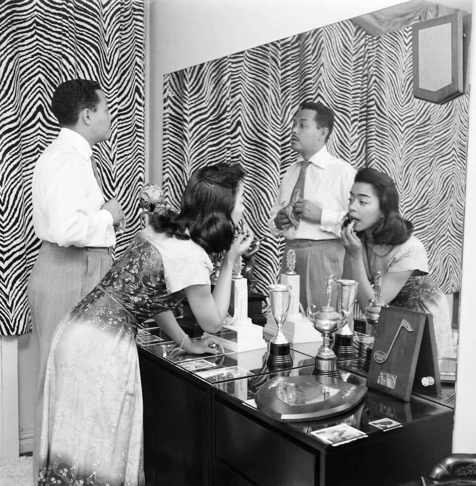 一男一女穿著1950年代服裝，在鏡子前化妝，梳妝台上放著獎盃。