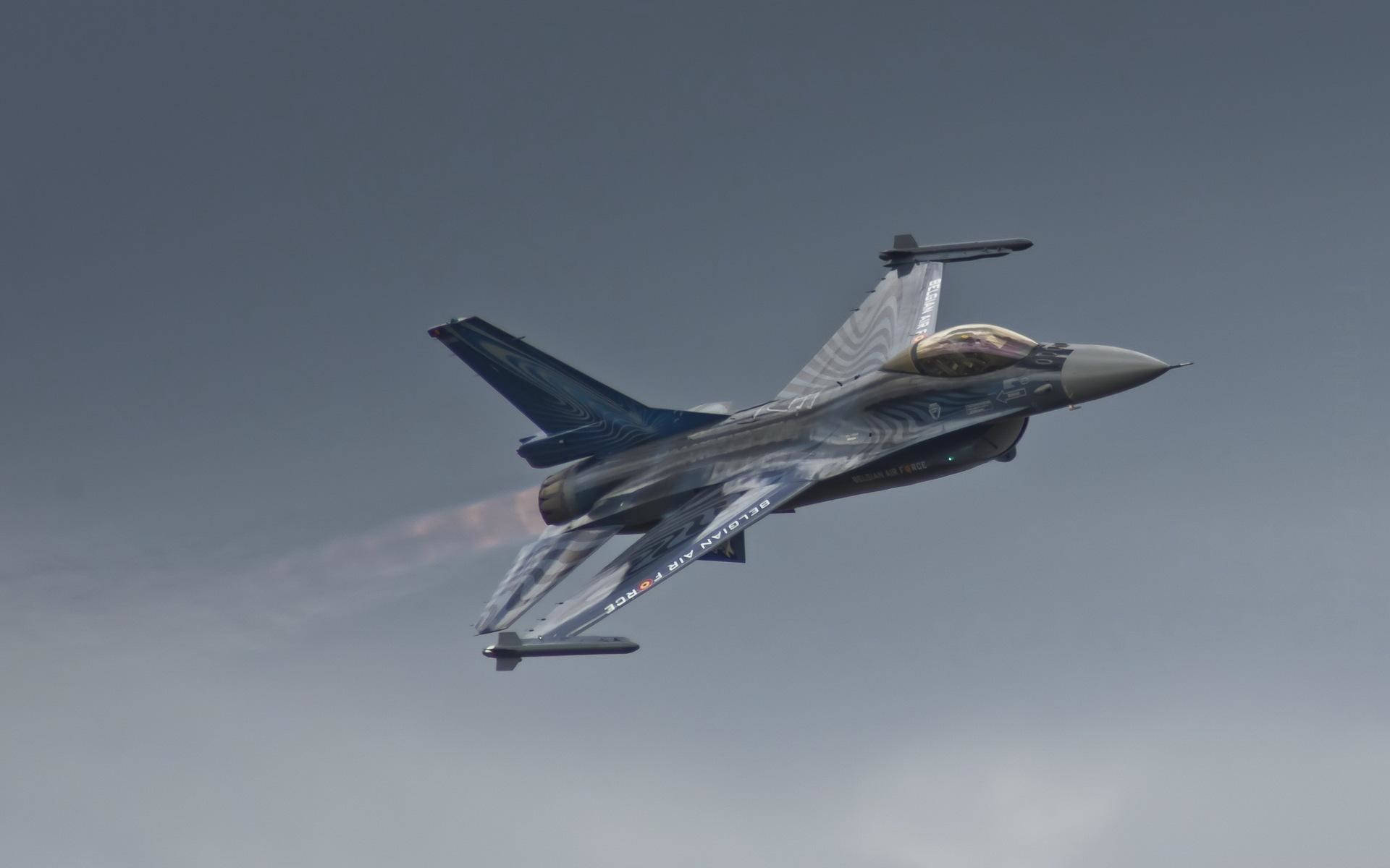 比利時空軍鷹隼 壁紙 — 一架流線型戰鬥機帶有藍色和灰色迷彩條紋，在雲層密佈的天空中翱翔，留下熾熱的排氣尾跡。